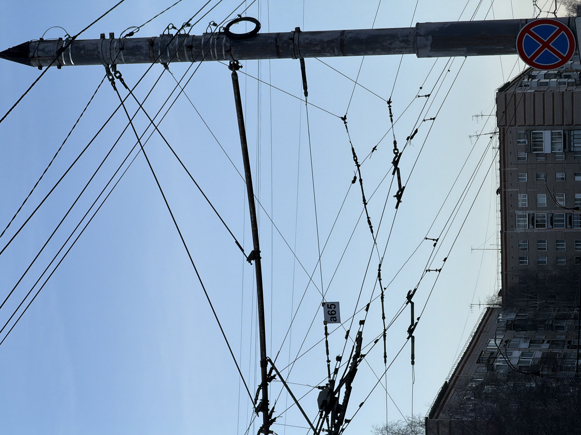 The image shows a view of overhead electrical lines and infrastructure in Izhevsk, Russia. The infrastructure consists of a horizontal pipe supported by vertical poles, with numerous wires and connections branching out. A "no parking" sign is attached to the pipe. The sign is round with a red border, a blue background, and a white "X" through the center. Behind the infrastructure, a multi-story concrete apartment building is visible with several windows. The sky is a pale blue, suggesting a winter day. The infrastructure has a tag on it which reads "a65".
