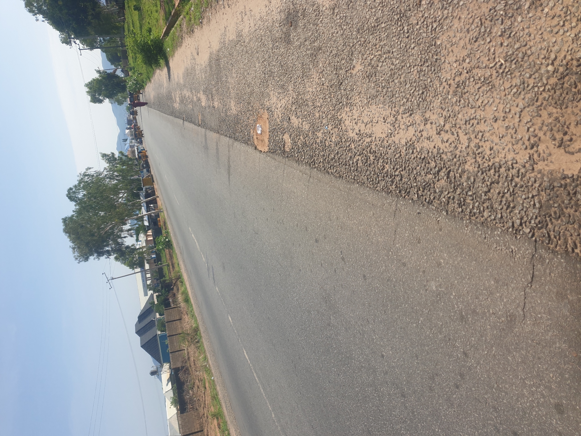 The image depicts a two-lane asphalt road in Jalingo, Nigeria, under a clear blue sky. The road is bordered by a gravel and dirt shoulder on the right, with sparse vegetation and some debris. On the left side of the road, there are buildings, including what appears to be a residential structure with a dark, gabled roof and a water tower, as well as other commercial or industrial buildings with flat roofs. Several trees with light-colored foliage line the left side of the road. In the distance, the road curves and shows traffic, including a few yellow tricycles (keke napeps) and a person walking along the shoulder. The overall scene is consistent with a road in a developing urban or semi-urban area.