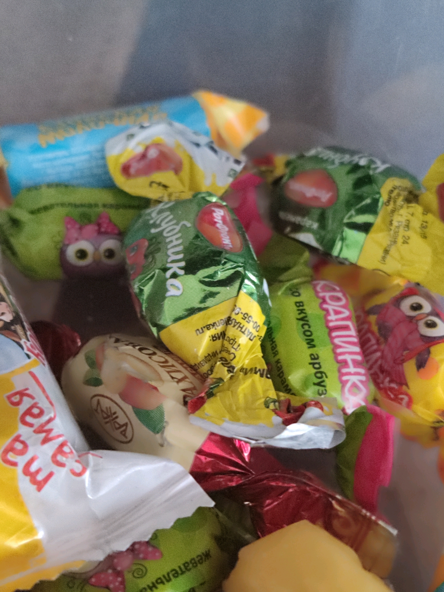A close-up, overhead shot captures a jumble of colorful, individually wrapped candies. The candies are primarily in shades of green, yellow, red, and blue, with some wrappers featuring cartoonish designs of owls and strawberries. The wrappers are mostly crinkled and layered on top of each other, suggesting they are in a container or pile. Some wrappers have text in Cyrillic script. The lighting is soft, and the background is a muted gray. No specific location in a city is discernible.