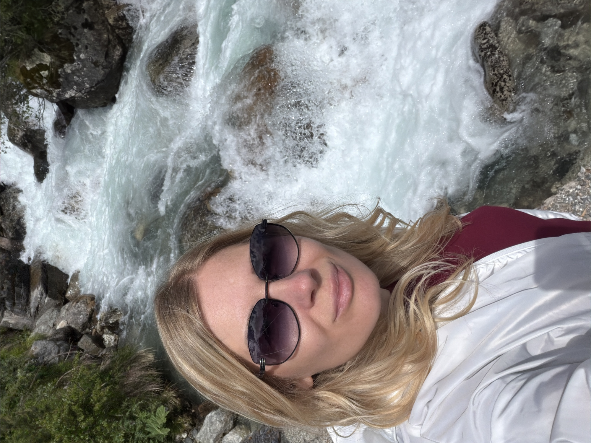 A woman with blonde hair and sunglasses takes a selfie in front of a rushing river in a rocky, mountainous landscape. She is wearing a white jacket over a maroon shirt. The water is turbulent and white, with large rocks visible beneath the surface and along the riverbank. Green vegetation grows on the rocky slopes to the left of the river. The sun is shining, casting bright light on the scene. The location is specified as Verkhne-Teberdinskoye rural settlement, Russia.