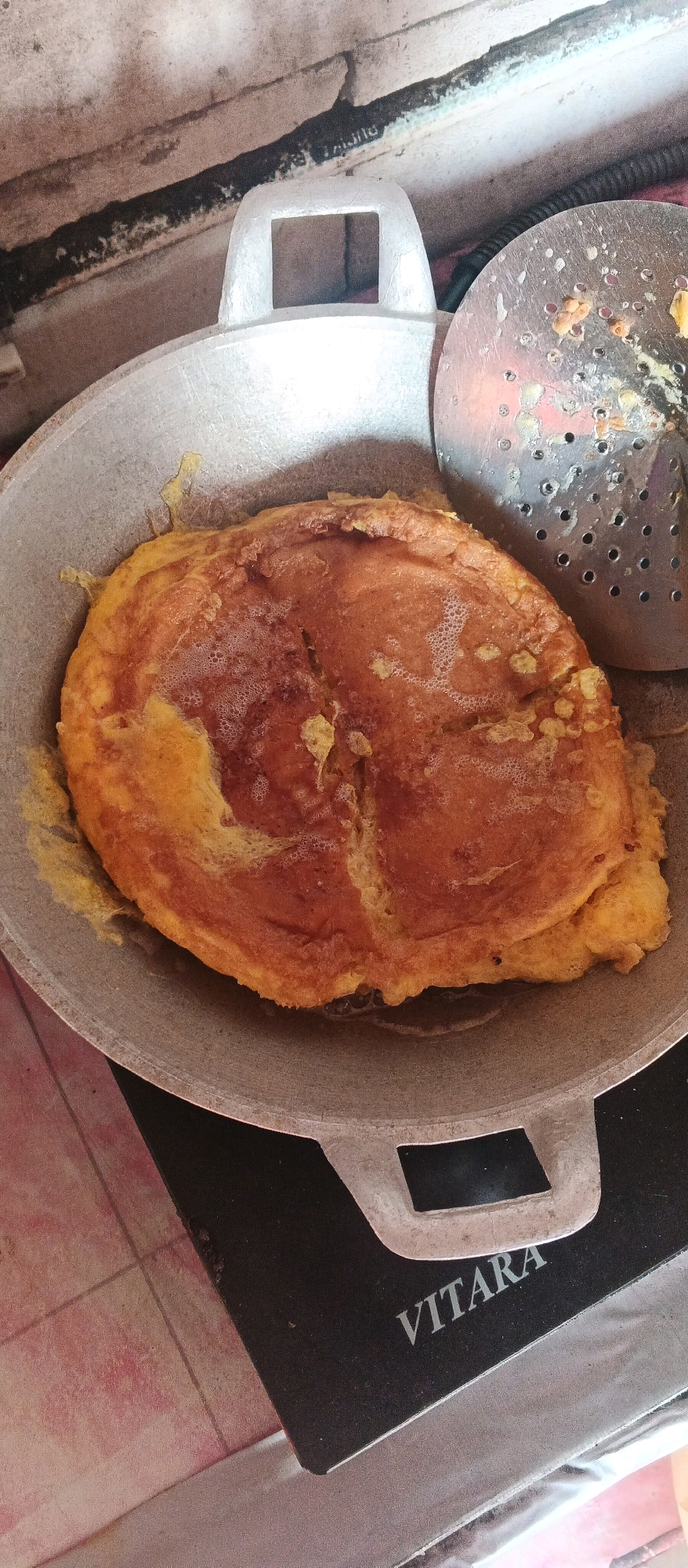 An overhead, close-up shot shows a golden-brown omelet cooking in a silver-colored wok. The omelet is round with irregular edges and has a slightly puffed surface with some air bubbles. A metal strainer with many holes, also with bits of food on it, is positioned to the right of the wok. The wok is resting on a black electric stovetop with the brand name "VITARA" visible in white lettering on its surface. The background appears to be a countertop or table with a textured surface, and possibly some tiles visible on the left. The overall lighting suggests an indoor setting. The image was taken in Bocek, Indonesia.