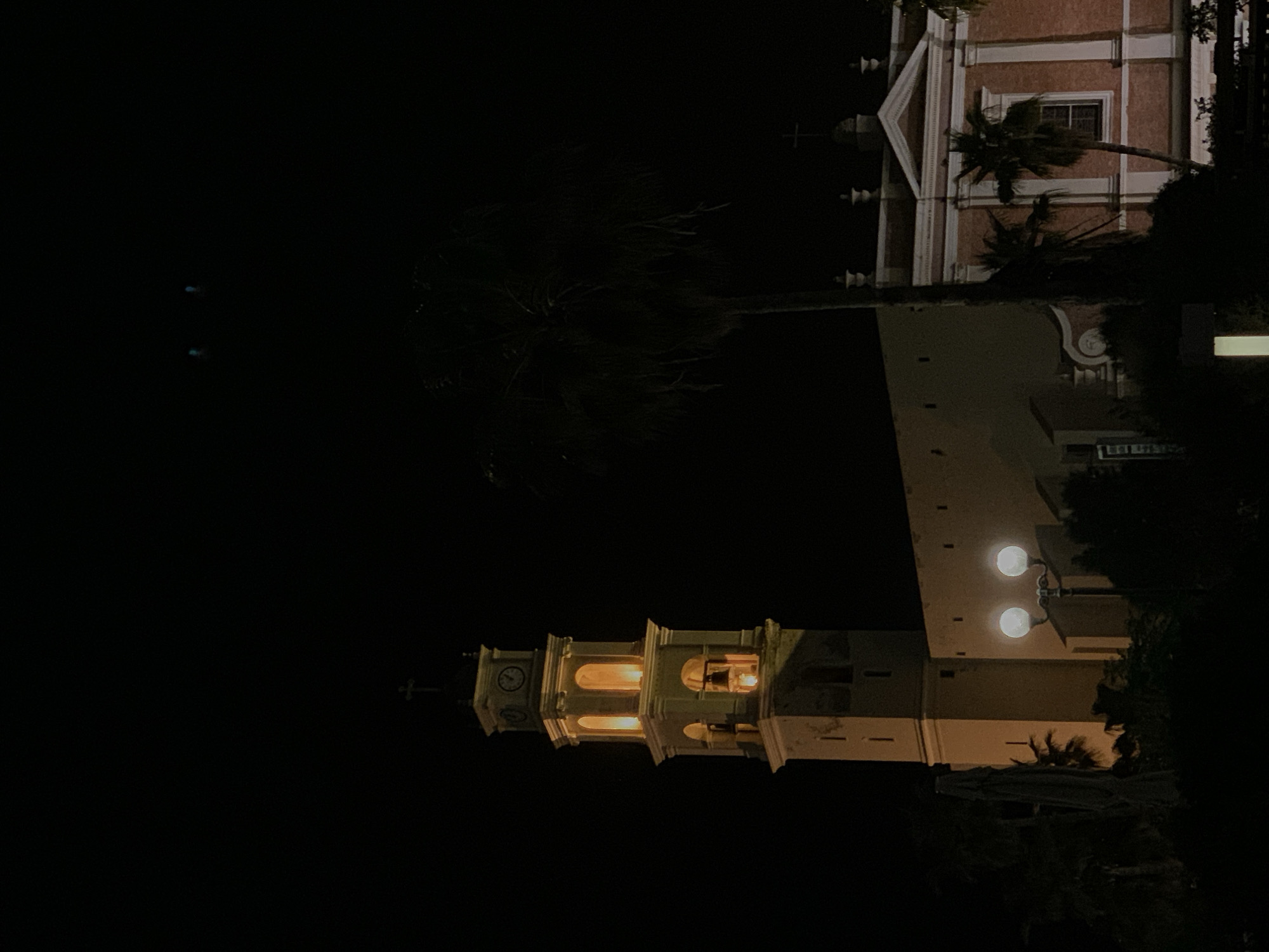 The image depicts a nighttime scene of St. Peter's Church in Jaffa, Tel Aviv, Israel. The church's clock tower is visible in the foreground, illuminated and showing its architectural details. Adjacent to the tower is another building, possibly part of the church complex, featuring a red facade with white trim. A tall palm tree is positioned between the two buildings. The rest of the image is predominantly dark, emphasizing the church's illuminated structure against the night sky. Street lights can be seen.