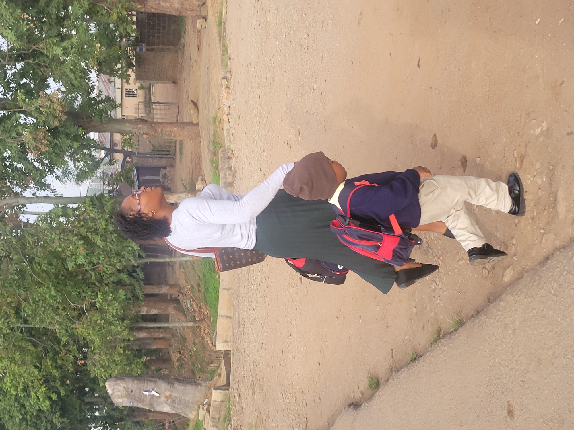 A woman and a child are walking on a dirt road near Jos, Nigeria. The woman has dark curly hair and is wearing glasses, a long-sleeved white shirt, and a long green skirt. She has a brown patterned purse over her shoulder. The child is wearing a school uniform including a brown hat, navy jacket with red trim, khaki pants, and black shoes. The child has a blue and red backpack. The road is dirt and gravel with patches of green grass. There are trees on the left side of the image, and buildings are visible in the background.