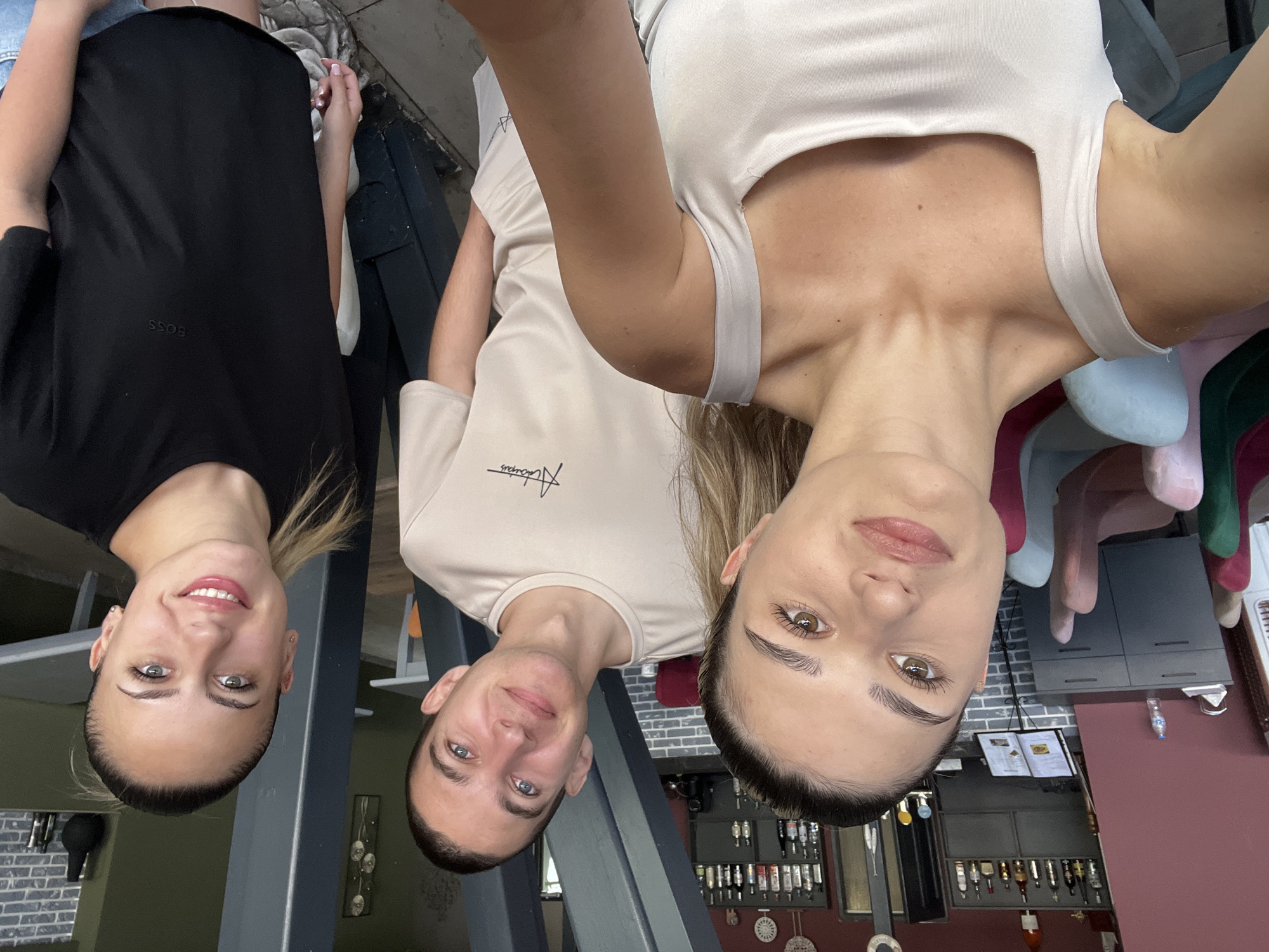 The media file shows a group of three young adults, two women and one man, posing for a selfie in what appears to be an indoor setting. The image is rotated 90 degrees counterclockwise.
The subject on the left is a woman with dark hair pulled back, wearing a black t-shirt with the "BOSS" logo visible. She is smiling broadly, displaying white teeth and a hint of dimples. Her eyes are a striking blue-green. To her right is a man with short brown hair and blue eyes, also smiling. He is wearing a light-colored t-shirt with some sort of graphic or text on it, possibly a logo. The third subject, on the far right, is a woman with long blonde hair. She is wearing a light-colored tank top. Her gaze is directed towards the camera, and she has expressive brown eyes and full lips.
The background suggests an indoor environment, possibly a cafe, restaurant, or bar. Visible elements include dark grey pillars, a brick wall, shelves stocked with bottles, and cabinetry. The lighting appears to be artificial, consistent with an indoor setting. The overall mood seems lighthearted and casual, suggested by the smiles and the selfie format. No specific time of day or weather conditions can be determined from the image. The location context provided is Lozenets, Bulgaria.
