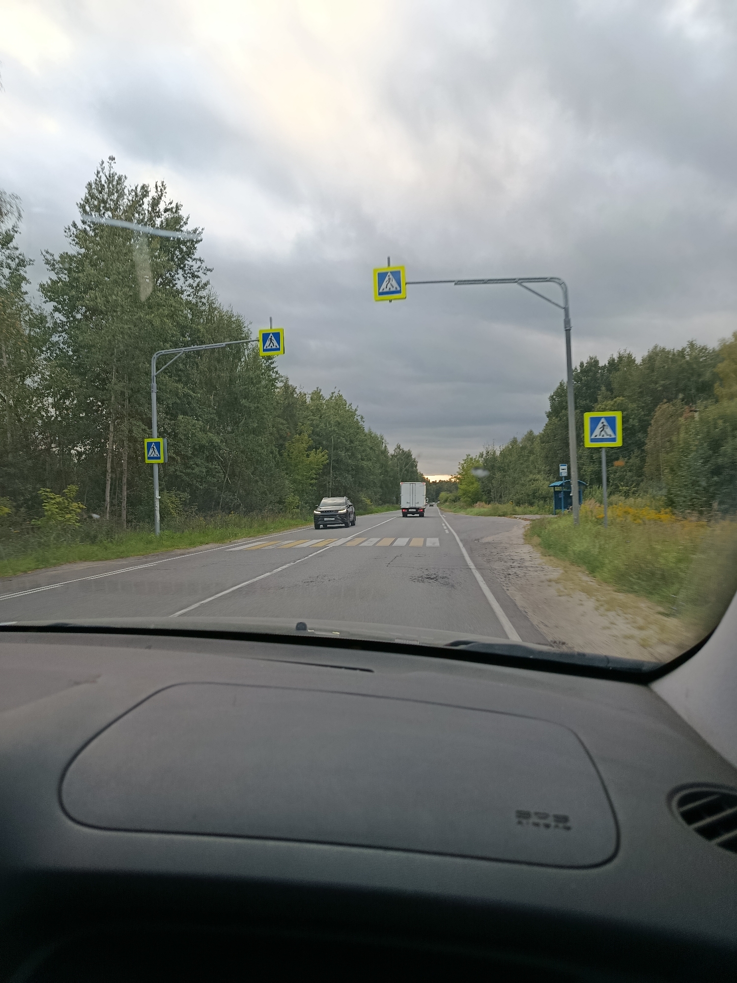 A view from inside a vehicle looking out at a two-lane road with pedestrian crossings. The road is bordered by a dense forest on both sides. The sky is overcast with grey clouds. There are multiple pedestrian crossing signs visible, featuring a yellow square with a blue border and a black silhouette of people crossing. One black SUV is visible driving on the road, and a white truck is driving in the opposite direction. The dashboard of the vehicle is visible in the foreground. The scene appears to be in or near the city of Vereya, Russia, indicated by the presence of these road signs and typical Russian landscape.