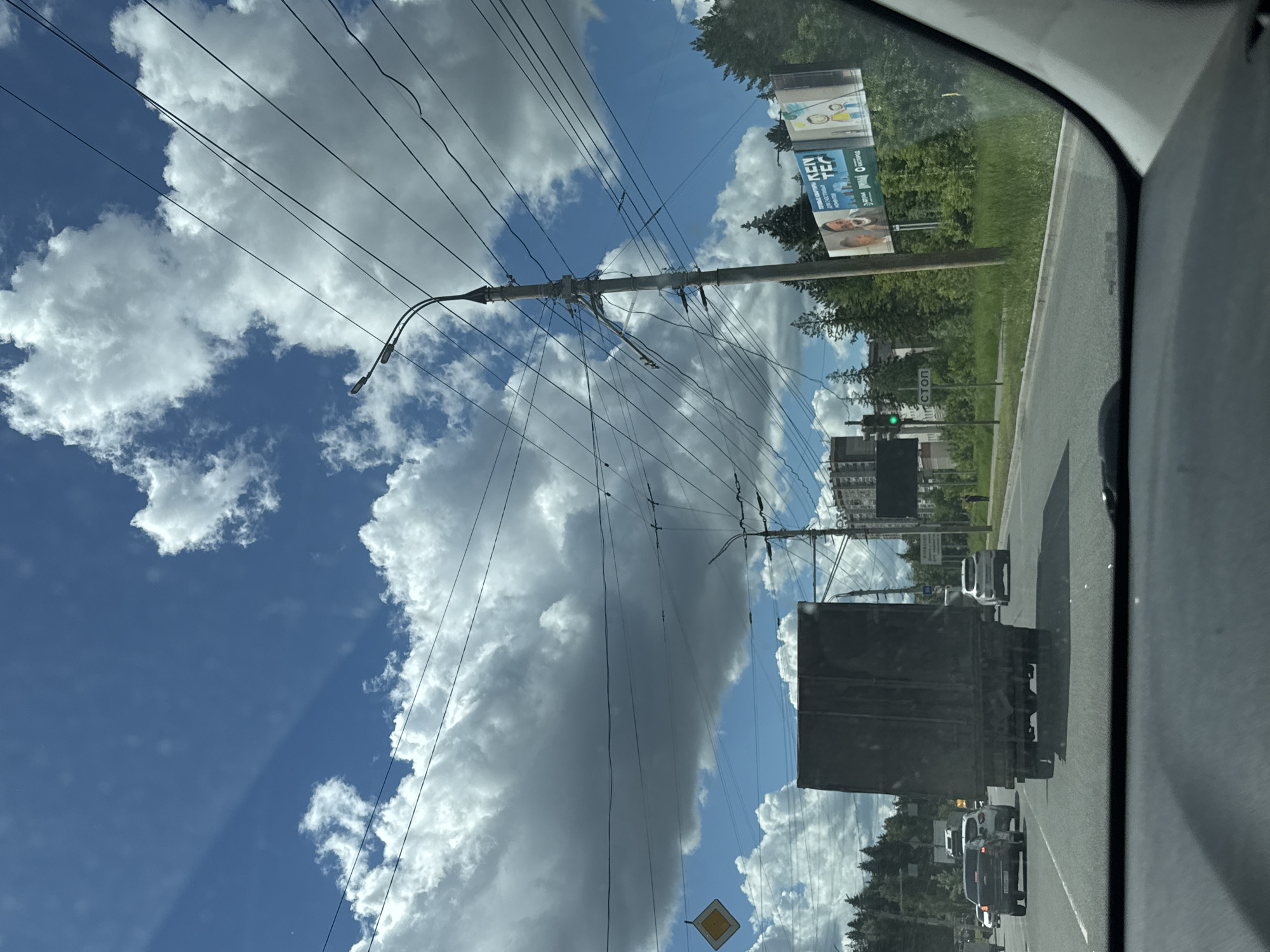 The image, taken from inside a vehicle, shows a street scene in Izhevsk, Russia. The sky is visible above with a mix of blue sky and fluffy white clouds. Utility poles and power lines run across the sky. On the right side of the image, a road is visible with cars and a truck. There are buildings and trees in the background. A billboard is on the side of the road. A yellow diamond-shaped road sign is also visible.