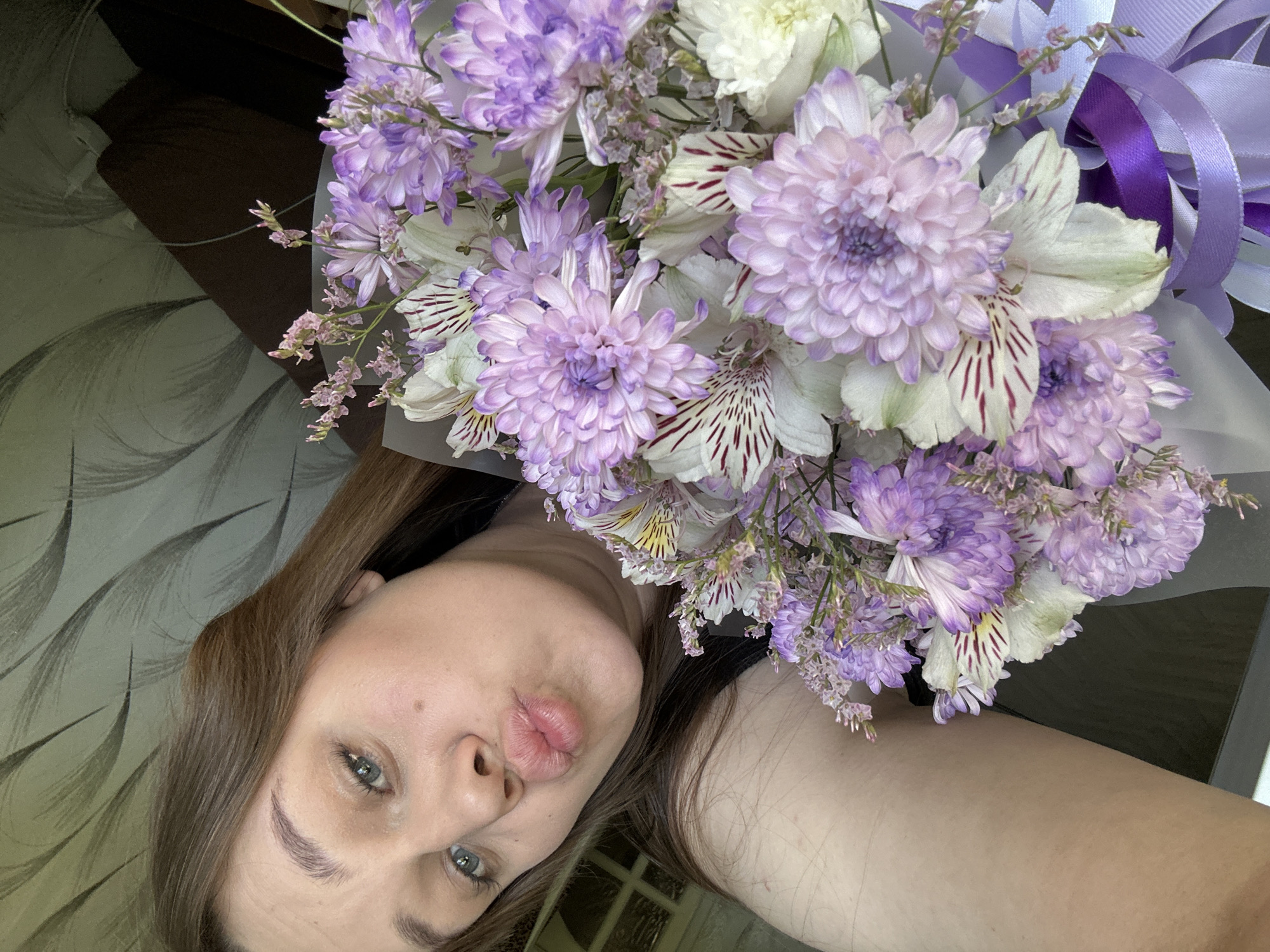 A woman with long brown hair is holding a bouquet of flowers in front of her face, pouting her lips towards the camera. The bouquet is a mix of purple and white flowers, including chrysanthemums and alstroemeria, and is wrapped with a white sheer material and purple ribbons. The background is a wall with a muted gray and brown pattern. The location is assumed to be Chaikovsky, Russia.