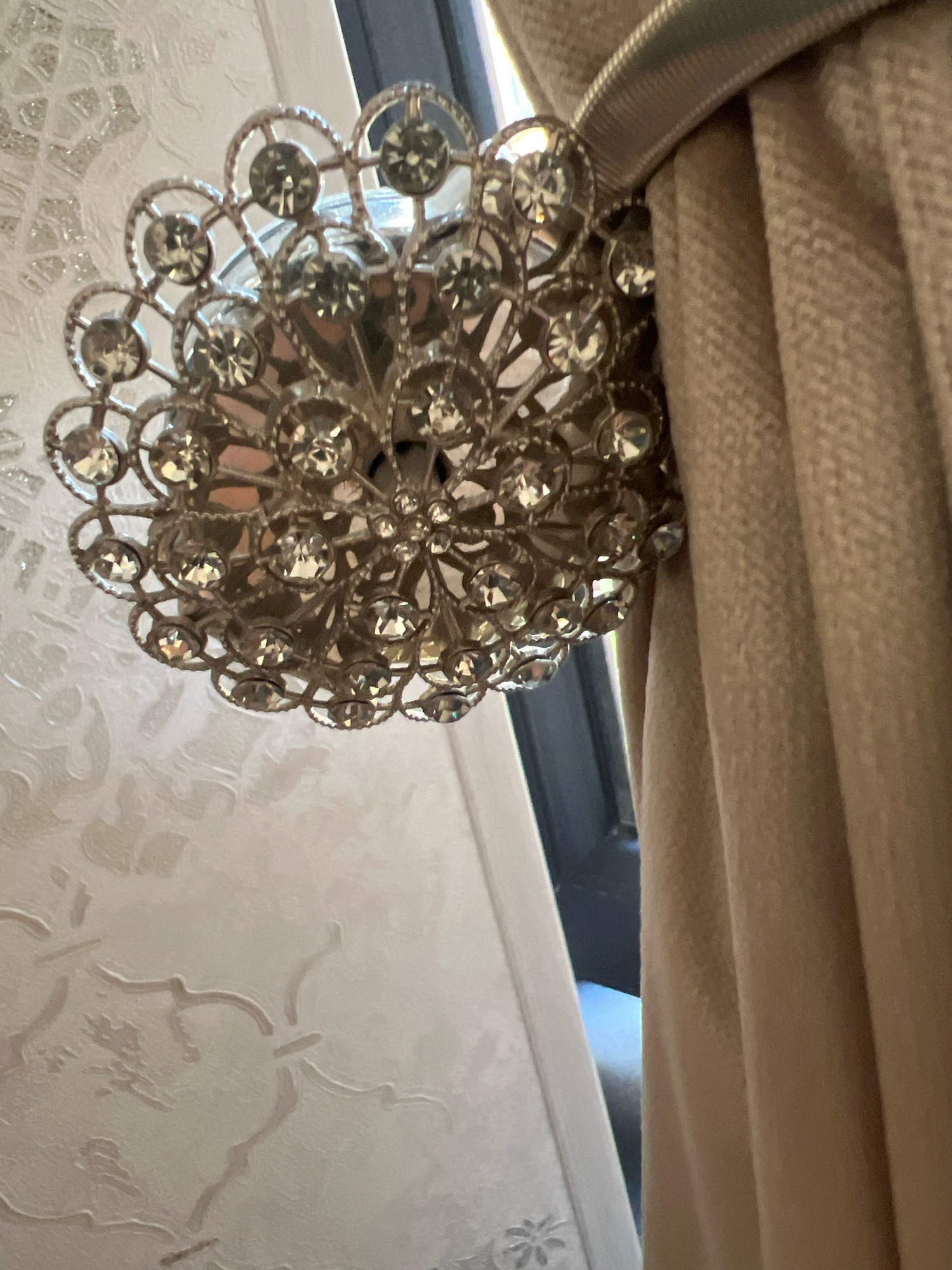 A decorative, metallic curtain tie-back adorned with clear, faceted stones is attached to a beige curtain. The tie-back has a circular, openwork design resembling a floral or starburst pattern. The curtain hangs vertically to the right of the tie-back. To the left of the tie-back, a textured white wall with a raised, ornate pattern is visible. The overall scene appears to be an interior setting, possibly a room in a residence.