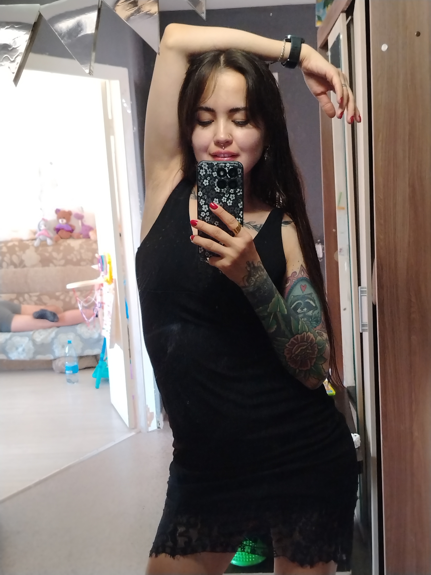 The image depicts a young woman in a black dress taking a mirror selfie. She has long, dark hair and is wearing a septum ring and a bracelet. Her left arm is raised, showcasing a full sleeve tattoo. The background appears to be an indoor setting, possibly a bedroom or dressing area, with a wardrobe on the right and a mirror on the left. In the reflection of the mirror, a couch with stuffed animals and a person lying down can be seen. There is also a bottle of water on the floor. The location of Noviy, Russia, is not visible or implied in the image.