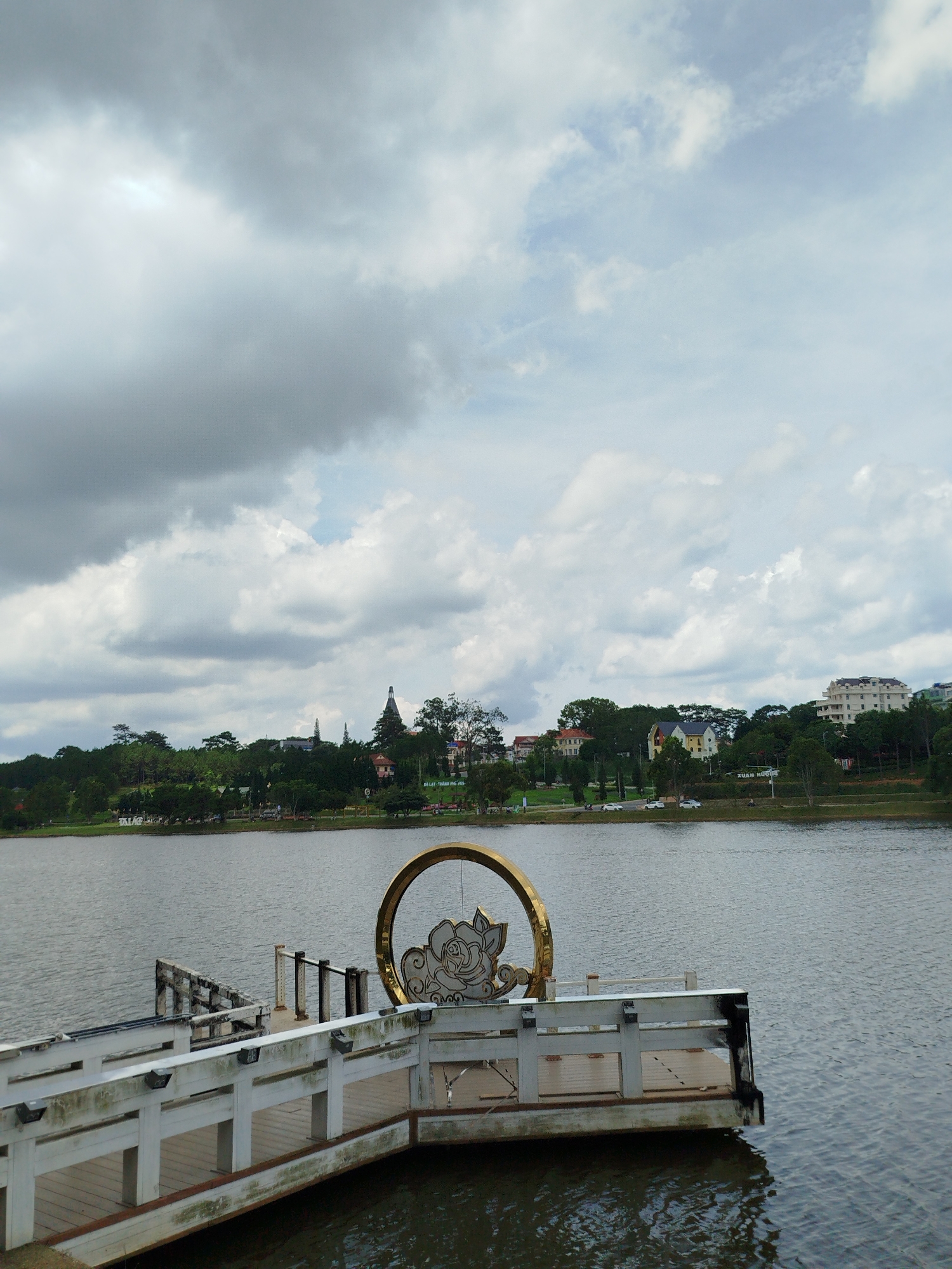 The image shows a wide shot of Xuan Huong Lake in Da Lat, Vietnam. In the foreground, a white wooden pier extends into the water. A large, golden circular art installation with a floral design is positioned on the pier. The lake is calm and reflects the cloudy sky. In the background, across the lake, a tree-lined shore is visible, with buildings and a church spire dotting the landscape. The sky is predominantly covered by dark, heavy clouds, suggesting an overcast day.