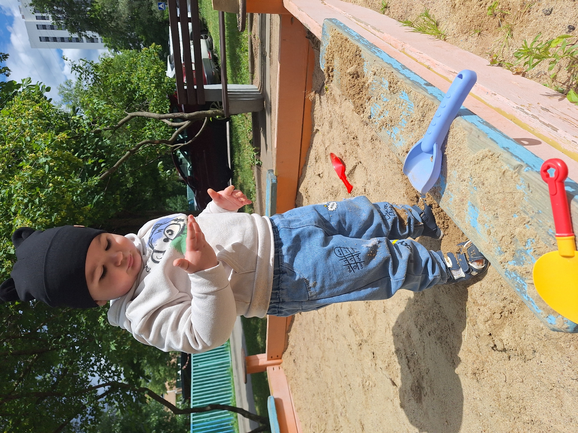 A young child is playing in a sandbox. The child, wearing a black hat with ears, a light-colored hooded sweatshirt, and blue jeans, is sitting in the sand. The child's hands are raised, as if in motion. The sandbox has wooden edges painted in pink and blue. A blue shovel and a red toy are in the sand near the child. A yellow shovel is also visible.
In the background, there are trees with green leaves, a building, and a park bench. The sky is blue with some clouds. The lighting suggests it is daytime. The overall scene is one of a child enjoying outdoor play. There is no visible text that can be read clearly.