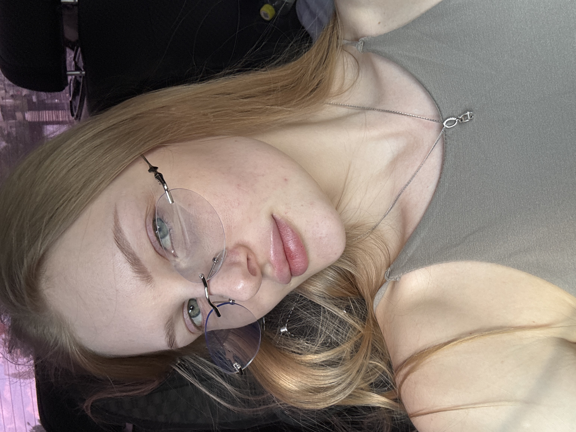 A fair-skinned young woman with light brown hair and blue eyes is depicted in this photograph. She is wearing rimless round glasses and a light gray top. A silver chain necklace with a small pendant is visible around her neck. The background is blurry, but appears to be indoors, and in the upper left corner there is a chair headrest. This photo appears to be taken from a close-up perspective. The picture does not provide information confirming or denying the location of this photo being in Perm, Russia.