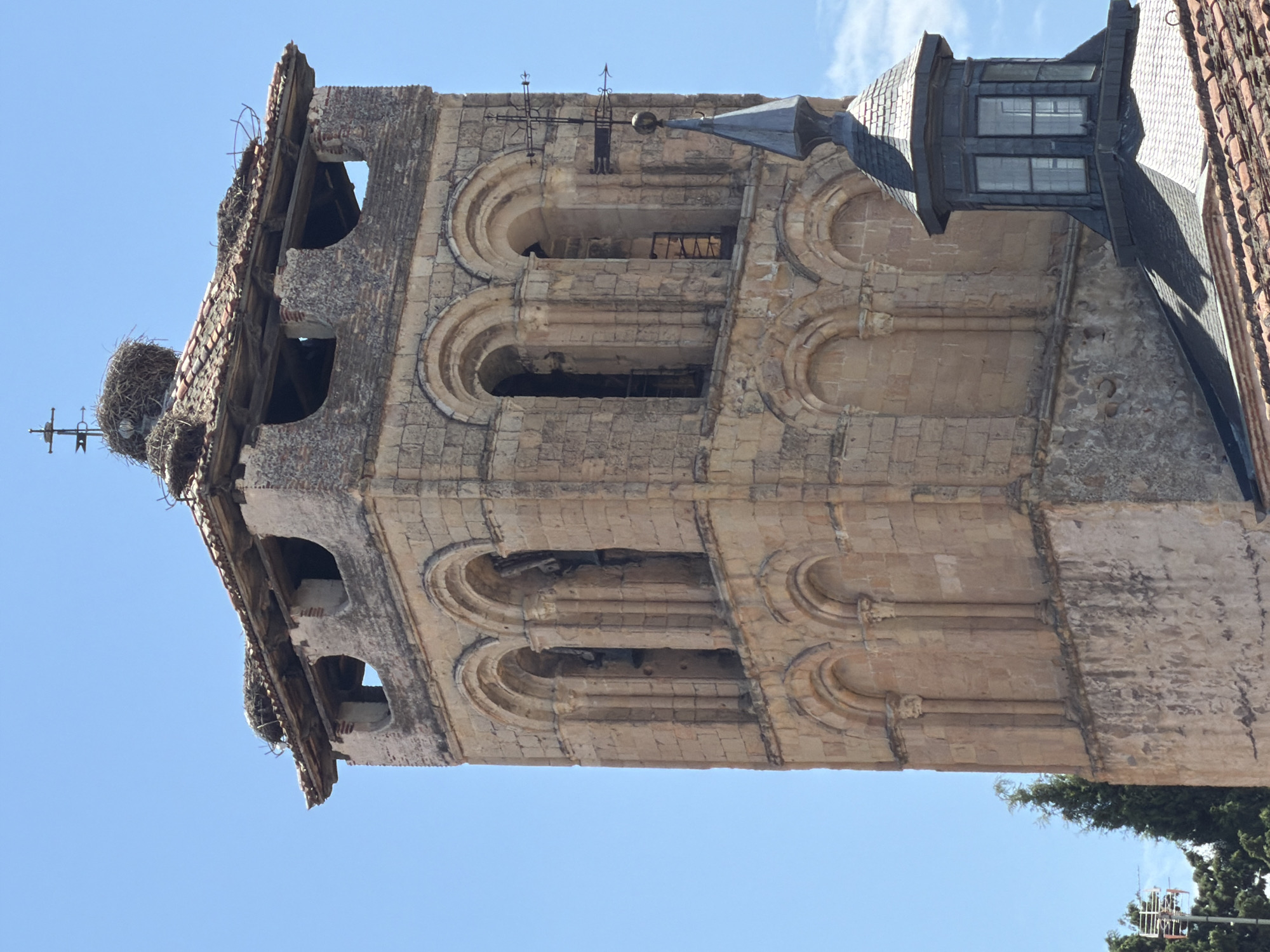 The image shows a close-up, low-angle view of the clock tower of the Cathedral of Segovia, Spain. The tower is constructed of weathered, light-colored stone blocks and features several levels of arched openings. At the very top of the tower, a large stork's nest is visible on the tiled roof. A cross stands behind the nest. A smaller, darker spire-like structure with windows is attached to the side of the tower, closer to the foreground and lower down, with its own tiled roof. The sky is clear and blue with a few wispy clouds. To the left of the tower, the tops of some trees and a small antenna structure are visible, suggesting an urban environment. The architectural style of the tower, with its Romanesque arches, is characteristic of medieval Spanish cathedrals.