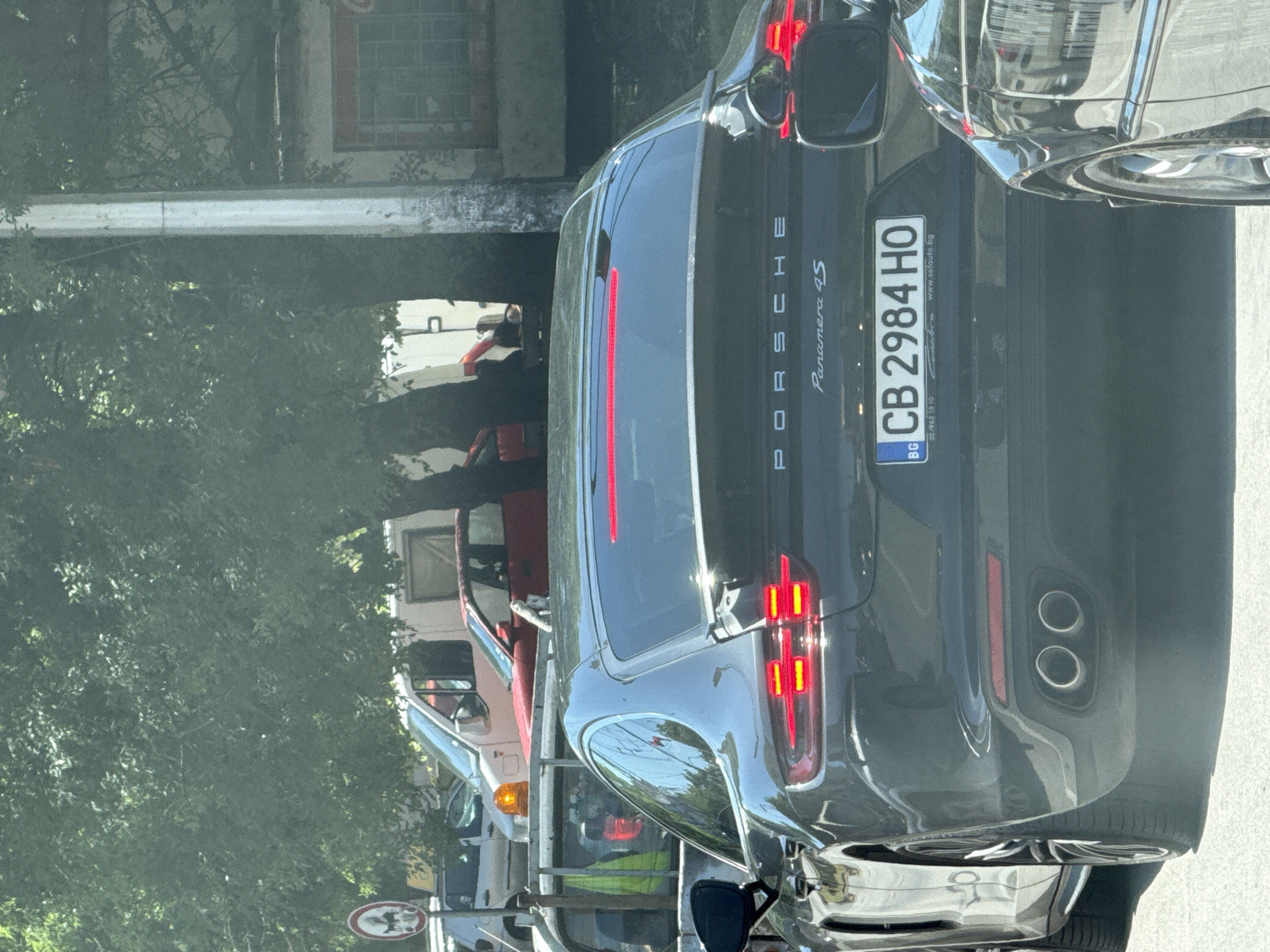 A dark gray Porsche Panamera 4S is stopped in traffic on a street in Pleven, Bulgaria. The license plate is CB 2984 HO. The car is in the foreground, with its rear facing the camera. The tail lights are illuminated. Other vehicles and trees are visible in the background. The environment appears to be an urban setting on a sunny day.