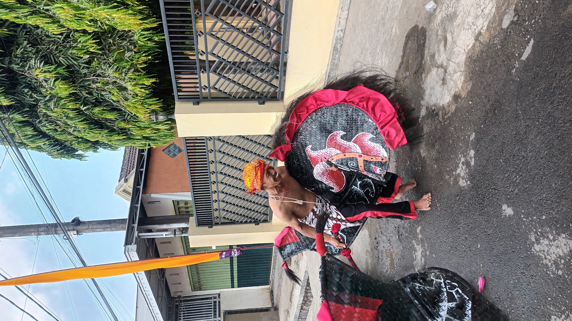 In Kras, Indonesia, a man is performing a traditional horse dance, known as Kuda Lumping, on an asphalt road. He is wearing a yellow and orange headdress and a patterned sarong, with his chest bare. The man is in motion, holding and manipulating a large, horse-like costume made of woven bamboo, fabric, and horsehair. The costume is predominantly black, adorned with red trim and a painted red and orange facial design. In the background, there are houses with tiled roofs, fences, and a tall green tree. Overhead, electrical wires are visible against a sky with scattered clouds. The road surface shows signs of recent rain, with puddles and damp patches.