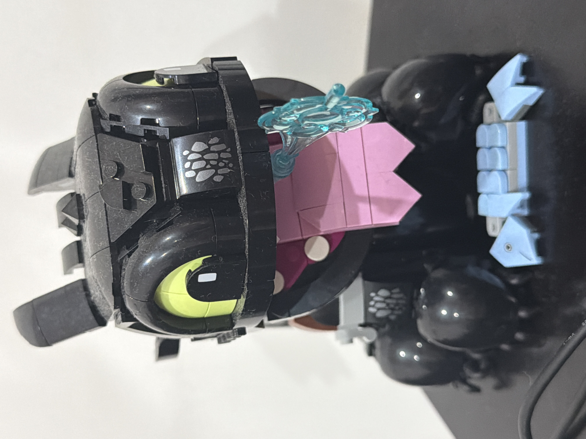 The image shows a Lego model of Toothless, a character from the "How to Train Your Dragon" franchise. The model is primarily black, with yellow eyes, light blue teeth, and a pink tongue. A translucent blue accessory is also visible. The model appears to be placed on a surface against a background that is partially white and partially black.