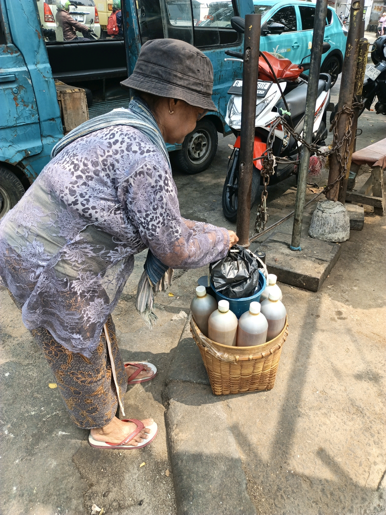 A woman wearing a patterned shirt, patterned pants, a bucket hat, and flip-flops is standing next to a blue truck and a motorcycle that is chained to metal poles. She is holding a black plastic bag over a basket filled with bottles of liquid. The scene appears to be on a street in South Jakarta, Indonesia, with buildings and other vehicles visible in the background.