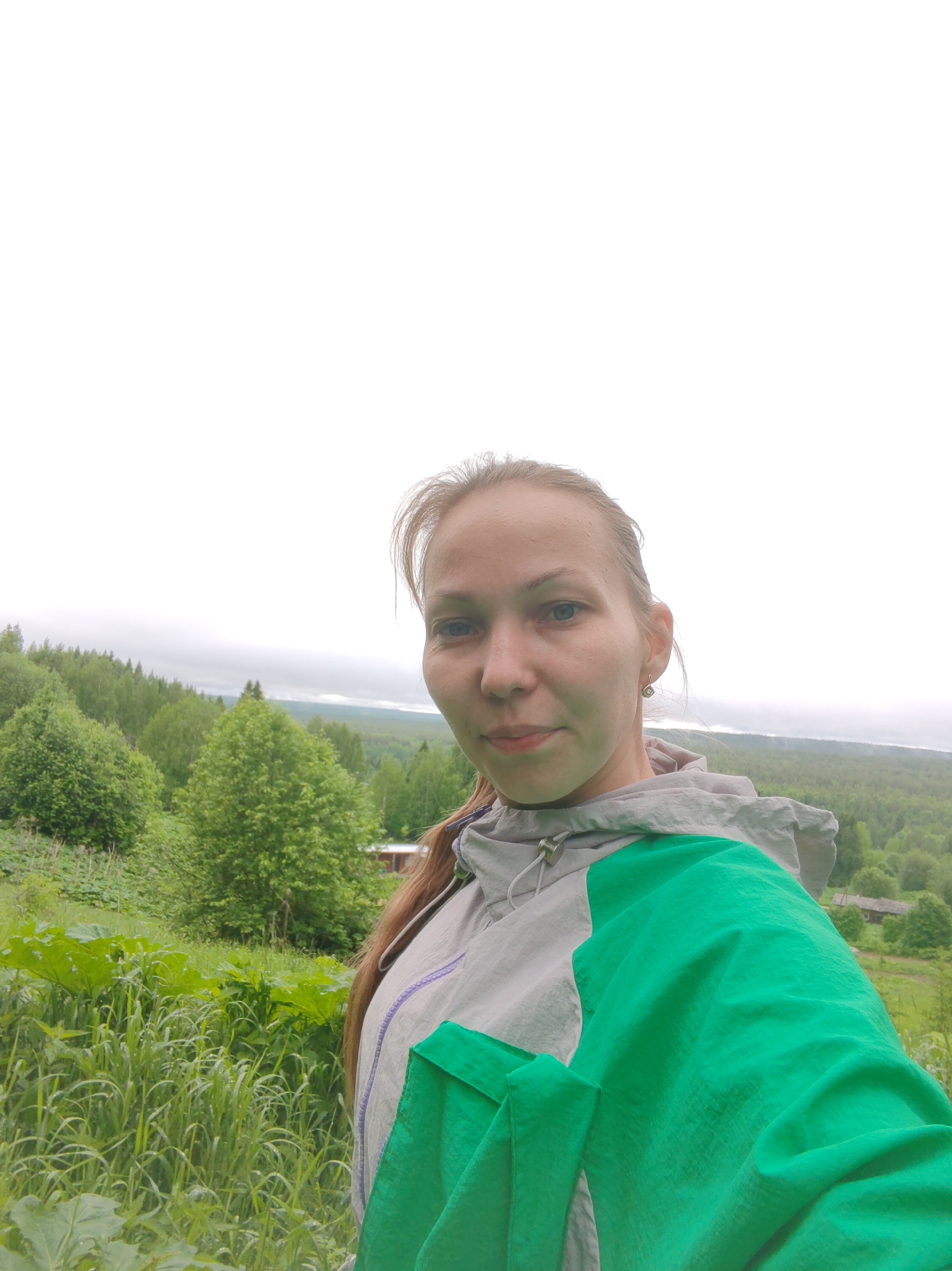 A woman is posing for a selfie outdoors in Чухлэм, Russia. She has fair skin, light brown hair pulled back, and is wearing a light gray jacket with a green sleeve. Behind her is a landscape of green trees, fields, and a cloudy sky. In the distance, a body of water is visible. Tall grass and vegetation are present in the foreground.