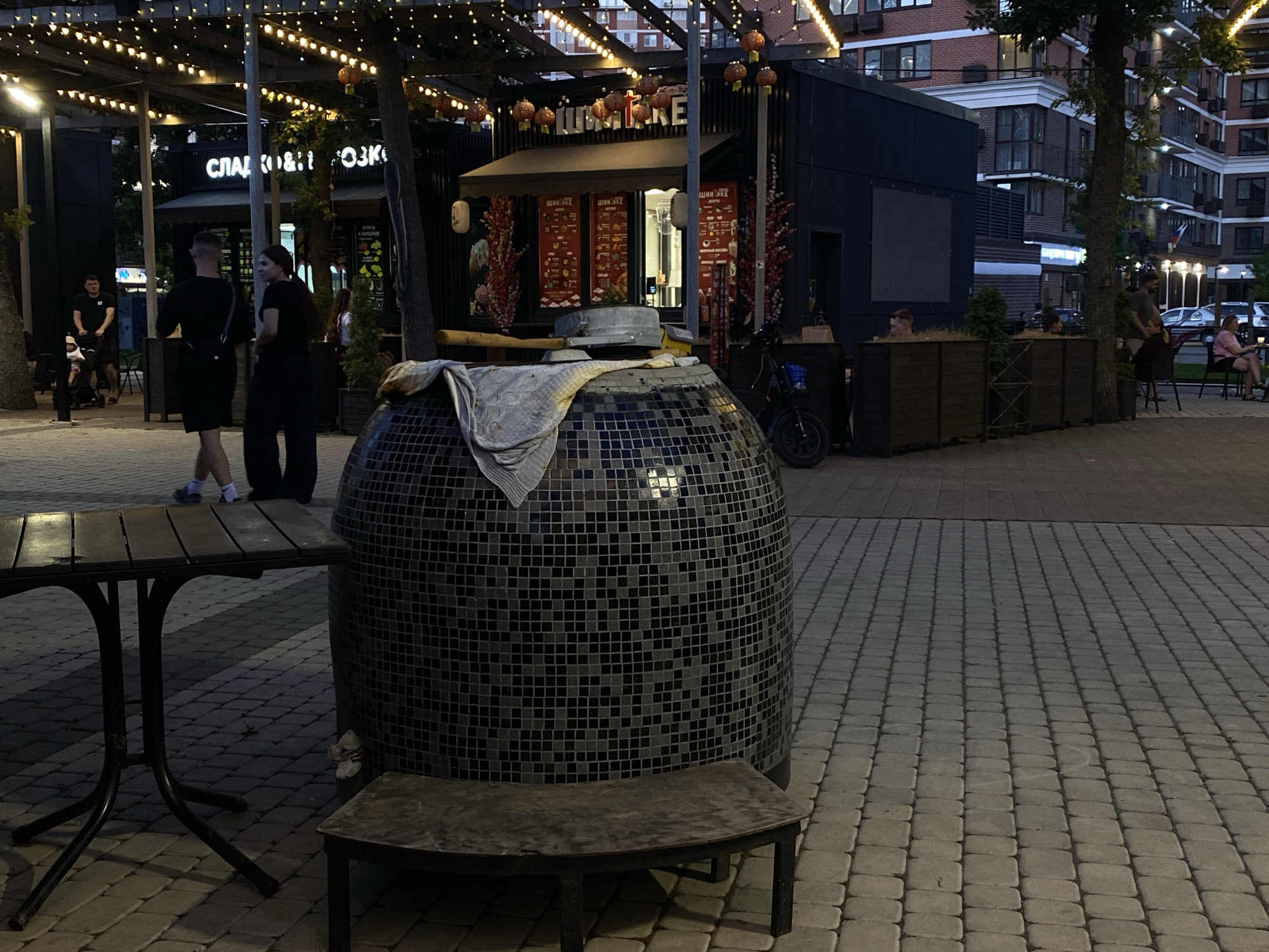The image depicts an outdoor seating area at dusk in Krasnodar, Russia. In the foreground, a dark metal table with a plank top stands on a patterned brick walkway. To the right of the table is a large, round, mosaic-tiled structure, possibly a tandoor oven, covered with a cloth. A dark, curved bench sits in front of the tiled structure.
Behind the table, two individuals, a man and a woman, are walking away from the camera. The man is wearing a black t-shirt and shorts, while the woman is in a black dress. Further back, a food stall with bright signage, likely advertising Asian cuisine, is visible. The stall features red lanterns and menu displays. A black electric scooter is parked to the right of the stall.
In the background, modern apartment buildings are illuminated. Several other individuals are seated at tables in the background, some engaging in conversations. The overall atmosphere suggests a commercial area with outdoor dining and entertainment. The lighting is a mix of natural twilight and artificial string lights illuminating the dining area.