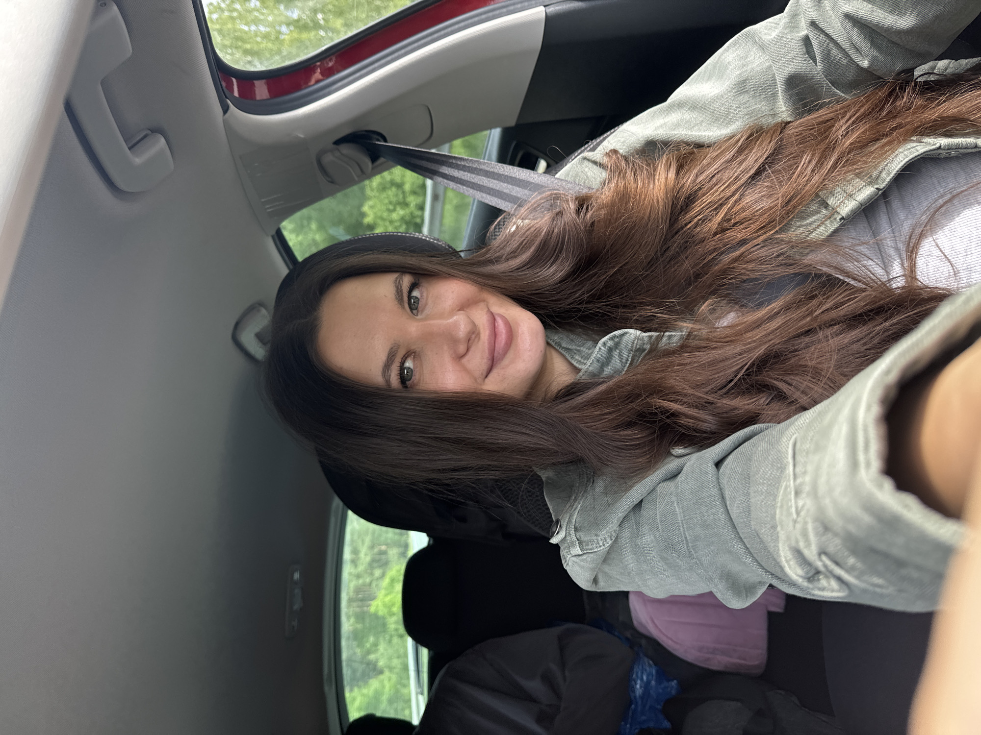 A woman with long, brown hair is sitting in the back seat of a red car. She is wearing a light green denim jacket and appears to be taking a selfie. The interior of the car is visible, including the headrest, seatbelt, and some items on the seat. Outside the car window, green trees can be seen. The location is Kremyonki, Russia.