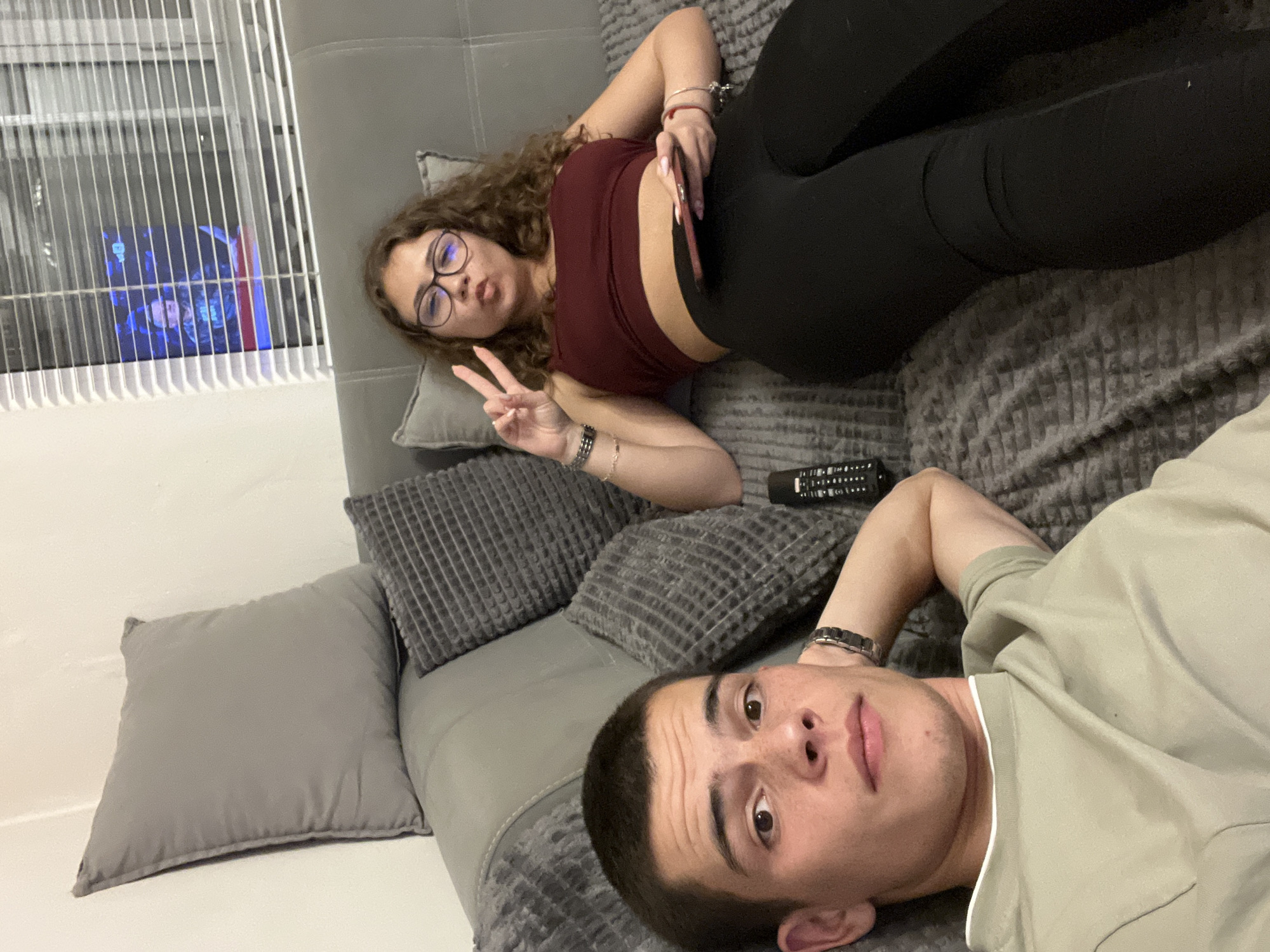 The image shows two people lying on a gray sofa in an interior space. The woman, wearing glasses, a maroon top, and black leggings, is positioned towards the upper-left of the frame and making a peace sign with her hand. She has curly hair and is holding a mobile phone. A remote control rests next to her arm. The man, wearing a light green t-shirt, is lying on the lower-right side of the frame. The location is not discernible as being in Plovdiv, Bulgaria; there are no obvious landmarks or signs to indicate this. The background includes window blinds and a wall. The environment appears to be a domestic living room.