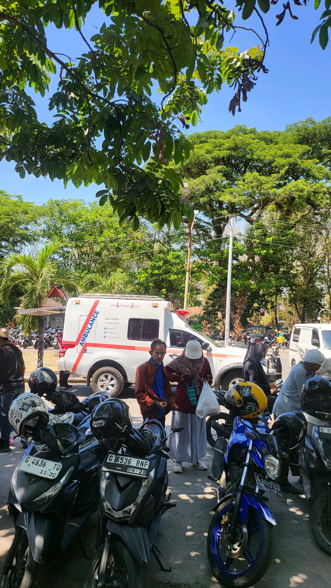 In Pakembaran, Indonesia, on a sunny day, an ambulance is parked near a group of people and motorcycles. The ambulance, predominantly white with red accents and the word "AMBULANCE" emblazoned on its side, has its doors closed. Several individuals, some in casual clothing and others wearing headscarves, are standing around the vehicle, suggesting they are either waiting for or assisting with something related to the ambulance. A line of parked motorcycles, mostly black and of varying models, fills the foreground and extends into the background, indicating a location where vehicles are commonly stored or gathered. The environment is characterized by lush green trees providing shade, with a clear blue sky overhead. The scene appears to be a public area, possibly a street or a parking lot within the city.