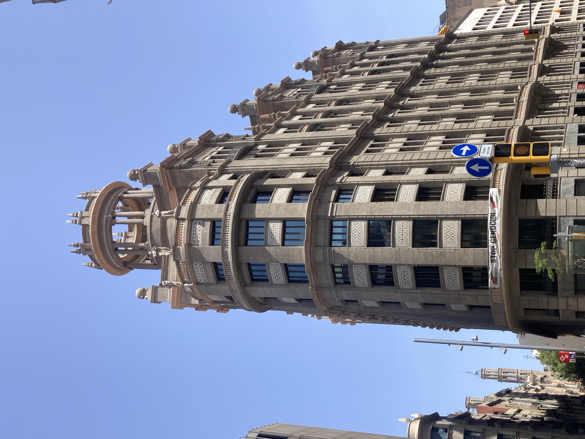 The image captures an upward view of the facade of a grand, ornate building in Barcelona, Spain, under a clear blue sky. The building is constructed of grey stone and features intricate decorative elements, including patterned friezes above windows and decorative statues on ledges. The upper portion of the building culminates in a circular, open-air rotunda supported by columns, topped with a decorative spire.
On the building's facade, a banner is visible, reading "STOP GENOCIDE." Below the banner, on the street level, a traffic light is present with red and yellow lights illuminated. There are also street signs, including a blue circular sign with a white arrow pointing right and another blue circular sign with a white arrow pointing up. A smaller sign below these has text in what appears to be Catalan, and a disabled access symbol is also visible.
To the left of the main building, another building's facade is partially visible, also appearing to be made of stone with multiple windows. Further in the background, a tall, historic bell tower with arched openings and a pointed roof rises above other buildings. The scene suggests a street in a bustling urban area, likely a commercial or historical district of Barcelona.