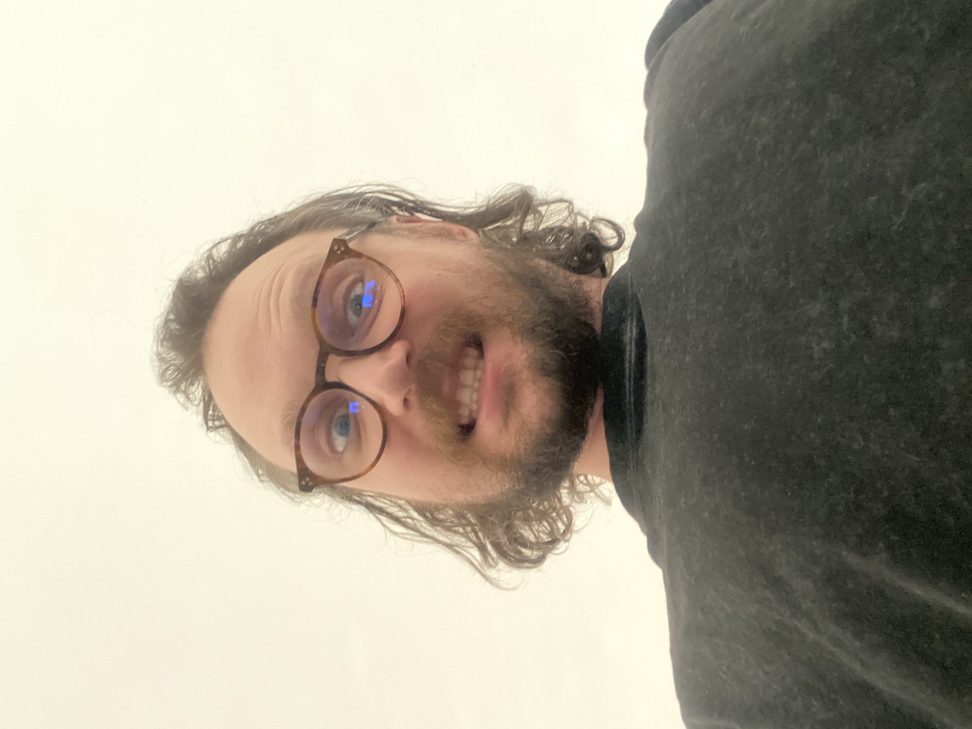 A close-up, rotated portrait shows a man with a beard, mustache, and dark, wavy hair. He wears brown-framed glasses, with bright blue-white reflections visible in the lenses. The image is rotated 90 degrees counter-clockwise, presenting his head tilted to the right. He looks directly at the camera with a wide, toothy smile, conveying a cheerful or amused expression. He is wearing a dark-colored (likely black or dark grey) t-shirt or top, visible on the right side of the frame. The background is a plain, light, off-white or very light beige color, suggesting an indoor setting with a minimalist backdrop. No specific location cues, time of day, weather details, or visible text are present within the image.