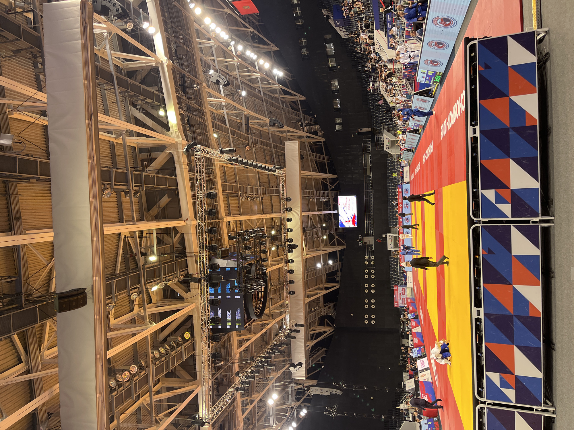 The image depicts an indoor judo competition taking place in the city of Krasnoyarsk, Russia. The scene is set within a large arena with a high, exposed ceiling featuring a complex network of beams, lighting rigs, and speakers. A large digital scoreboard is visible suspended from the ceiling, displaying competition information.
The judo mat is a prominent feature, divided into colored zones: a red outer area and a yellow inner area where the matches occur. Several athletes are present on the mat, some actively engaged in bouts and others waiting or walking. One judoka in a white gi is on the ground, being held by an opponent in a blue gi. Another athlete in a blue gi stands near the edge of the mat, observing. Officials or coaches in suits and judo uniforms are also visible on and around the mat.
Spectators are seated in tiered seating areas surrounding the competition floor, though they are largely out of focus. A decorative barrier with a geometric pattern in red, white, and blue is positioned in the foreground, separating the viewer from the judo mat. The overall environment suggests a professional sporting event.