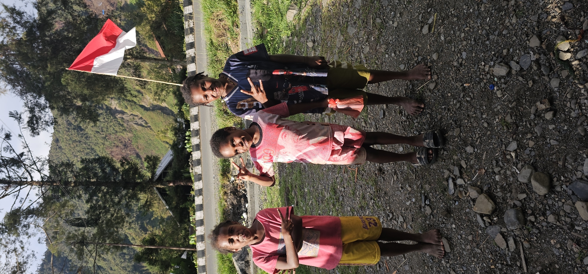 Three young girls, likely of Papuan descent, stand on a rocky ground in front of a paved road. The girl on the left wears a pink t-shirt and yellow shorts, her arms crossed, and she is making a peace sign with her fingers. The girl in the middle wears a pink t-shirt with a graphic print and has her hair styled in braids. She is also making a peace sign. The girl on the right wears a dark blue t-shirt with a soccer-themed graphic and is making a hand gesture that resembles horns. She has dark, curly hair. A red and white flag, likely the Indonesian flag, is visible in the background, planted on a wooden pole. The scene is set against a backdrop of lush green mountains and trees, with a partially visible building with a triangular roof in the distance. The lighting suggests it is daytime, possibly morning or afternoon, with sunlight casting shadows. The overall environment appears to be rural or semi-rural, with evidence of a road and some scattered debris on the ground. The location is specified as Puncak Jaya, Indonesia.