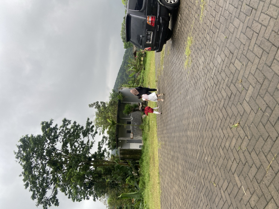 The image depicts a paved parking lot with brick pavers. A woman wearing a hijab and a long black top and black pants is walking away from the camera, followed by two children, a girl in a pink dress and a boy in a red shirt. The woman is in the foreground, with the two children behind her. The background is a low, white building with "Toilet" written on it. The building is adjacent to a tall tree. In the background is a black SUV parked on the edge of the parking lot. The SUV is facing the camera, with the back of the vehicle visible. The scene is in the location of Kecamatan Trawas, Indonesia at coordinates (latitude: -7.661, longitude: 112.62). The sky is overcast, with clouds covering the sun.