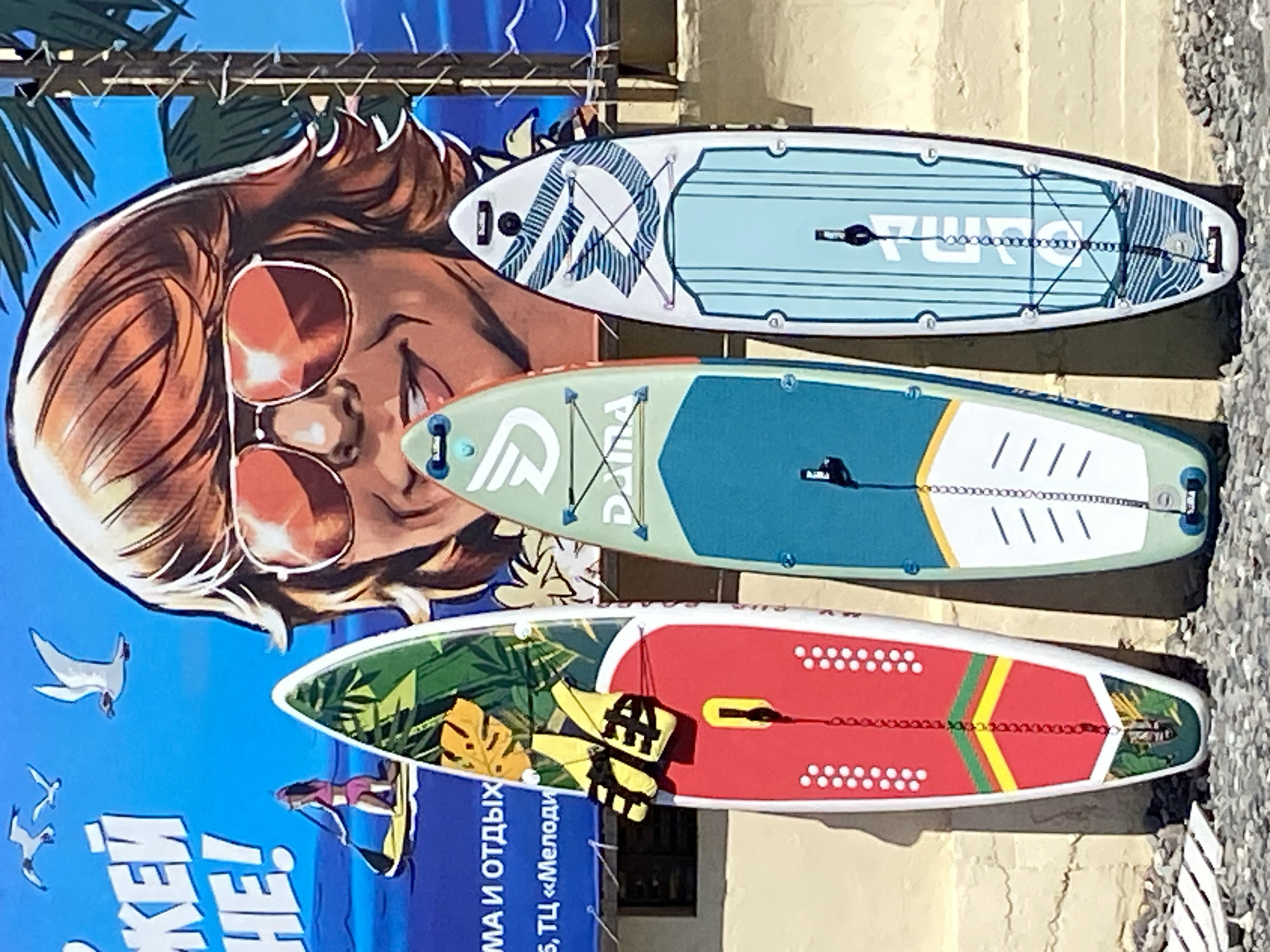 The image shows three stand-up paddleboards leaning against a concrete wall. The paddleboards are positioned vertically. The leftmost paddleboard has a red base with yellow and green stripes and a tropical leaf design at the bottom, with a pair of yellow life vests attached near the top. The middle paddleboard is light green with a white top section featuring a blue logo and white lines. The rightmost paddleboard is white with blue wave-like designs and a light blue section at the bottom. Behind the paddleboards is a large mural featuring a woman with blonde hair and sunglasses, and palm trees against a blue sky with seagulls. Text in Russian is visible on the mural, and some text is also visible on the paddleboards. The ground in front of the wall is covered with small stones and some white wooden planks. The overall setting appears to be an outdoor location, possibly near a beach or waterfront. The specific location in Sochi, Russia, is a wall with a mural and paddleboards, likely a commercial establishment or an advertising display.