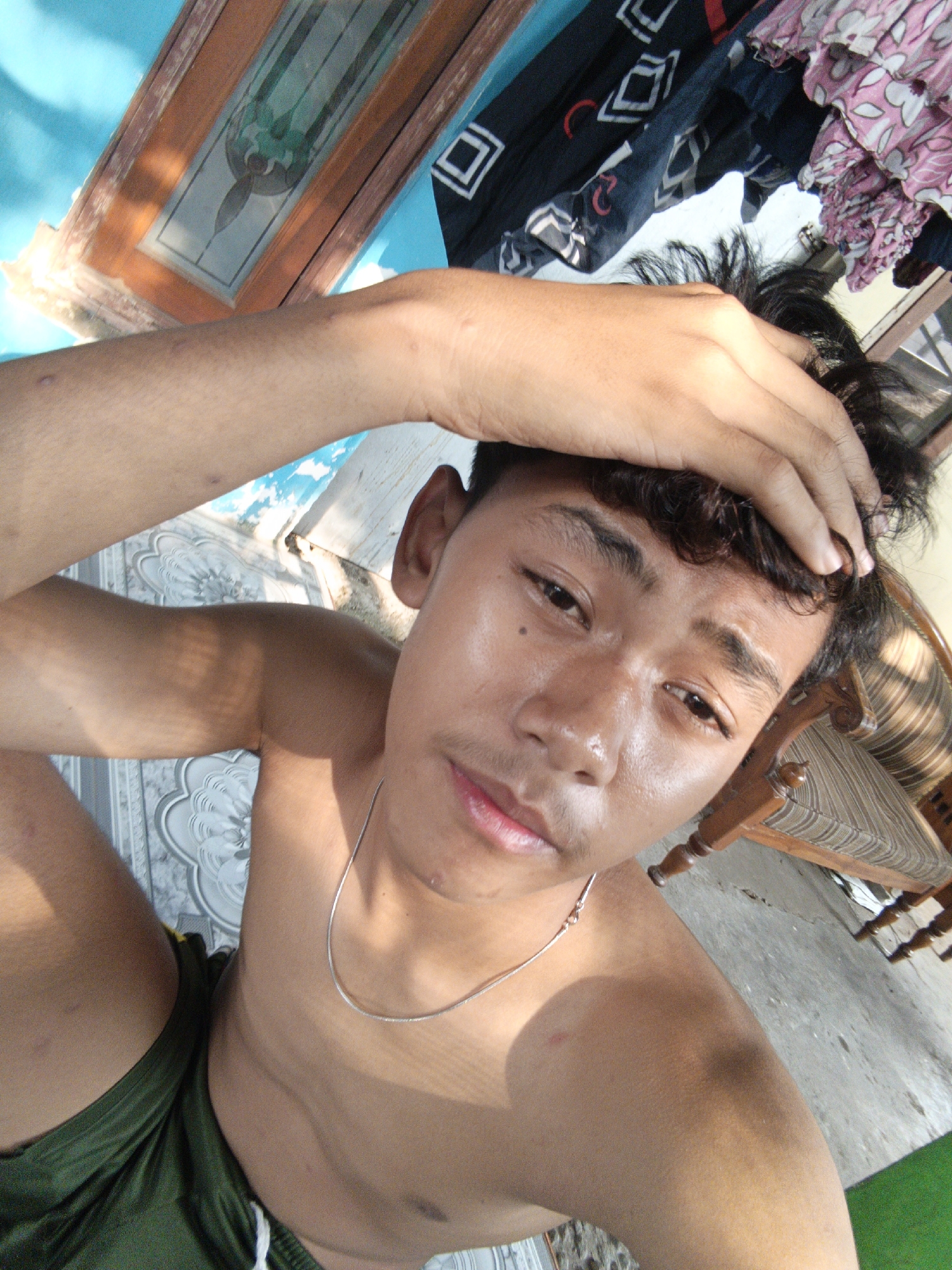 In the city of Temuwulan, Indonesia, a young male is captured in a selfie. He is wearing green shorts and a silver chain around his neck. His right arm is raised, with his hand resting on his head, obscuring a portion of his face. The background reveals a tiled floor, a wooden chair with striped upholstery, and what appears to be laundry draped over a railing. The scene is illuminated by natural light, casting shadows on the floor and the individual's body.