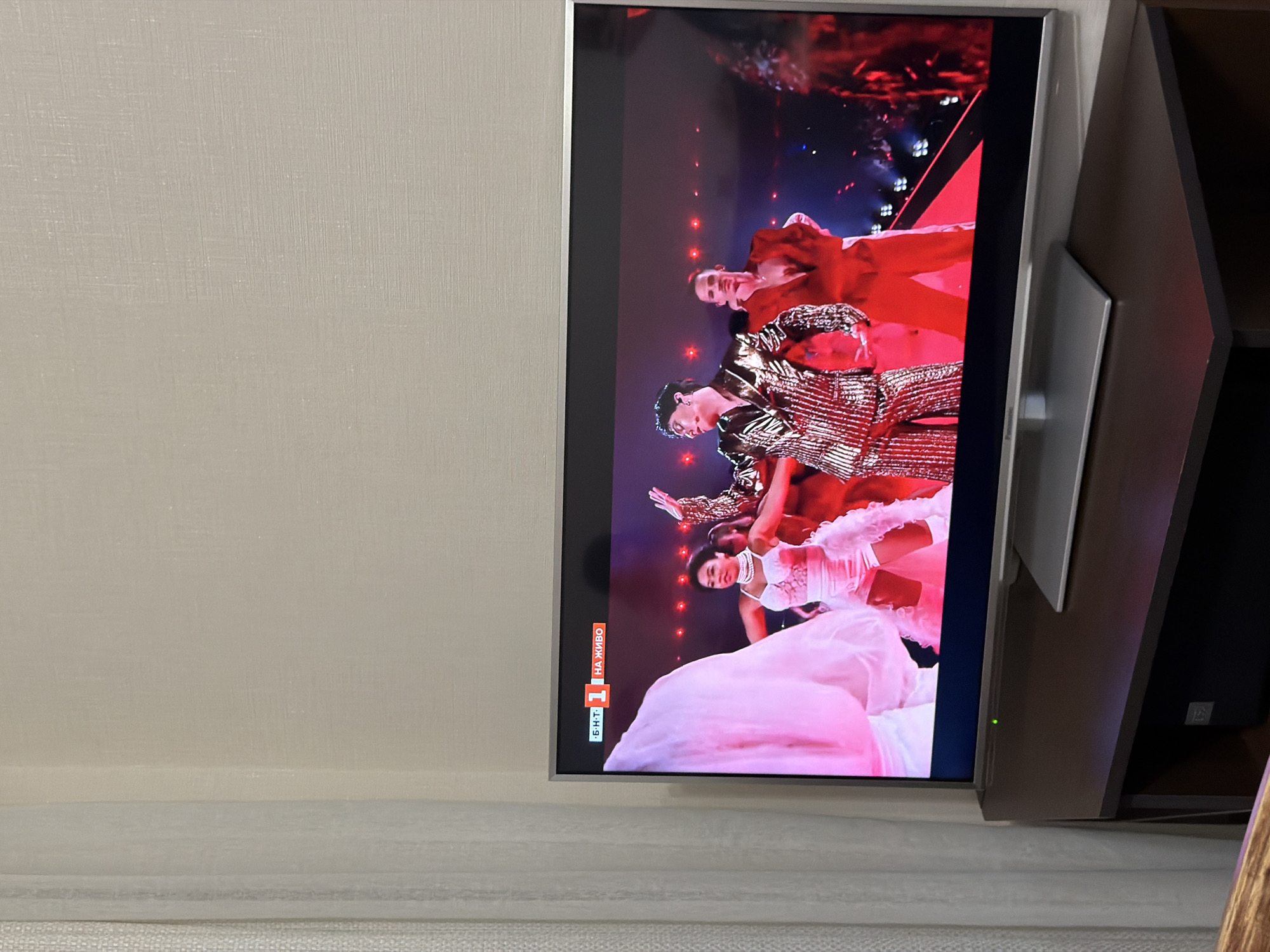 A television mounted on a wall displays a dance performance featuring four individuals. One woman is prominently wearing a large, light pink dress, while the others wear shiny, gold-colored outfits. A Bulgarian network logo ("БНТ 1 НА ЖИВО") is visible in the lower-left corner of the screen. The environment suggests a domestic interior setting. The location, Burgas, Bulgaria is not determinable from the image.
