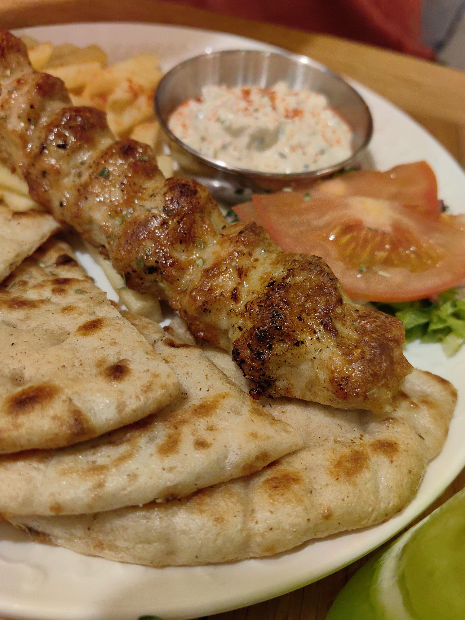 This forensic image captures a close-up of a prepared meal on a white, round plate, situated in Sochi, Russia. The primary subject is a single skewer of grilled meat, likely chicken, displaying a golden-brown, slightly charred exterior with visible seasoning (green herbs and dark specks). This skewer rests atop and alongside several pieces of lightly toasted flatbread, which exhibit browned spots. To the upper right of the skewer, a small, metallic ramekin holds a creamy white sauce, generously sprinkled with red flakes (likely paprika) and faint green herbs. Behind the skewer, partially blurred, are golden-yellow elements consistent with french fries or potato wedges. To the right, a side of salad includes a prominent slice of red tomato and green lettuce. A blurred green and white object, possibly a lime wedge, is visible at the bottom right. The background features a soft-focus wooden surface and a blurry reddish element, possibly fabric. No individuals are present in the frame.