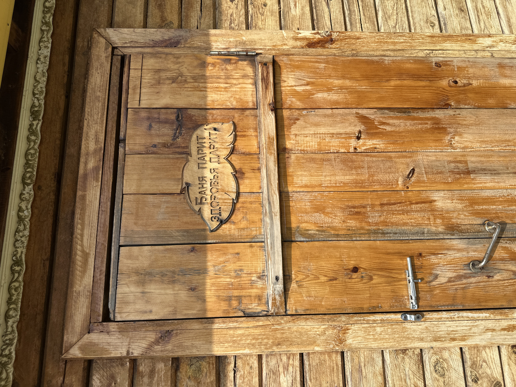 The image shows a wooden door of a sauna. The door is made of vertical planks of wood and has a metal bolt and handle. A decorative wooden sign with Cyrillic text is affixed to the upper part of the door. The sign is shaped like a cloud and reads "Баня парит, здоровья дарит" which translates to "The banya steams, health bestows". The door is set into a wooden wall, also made of horizontal planks. There is ornate trim above the door. The lighting suggests it is daytime, with sunlight casting shadows on the door.