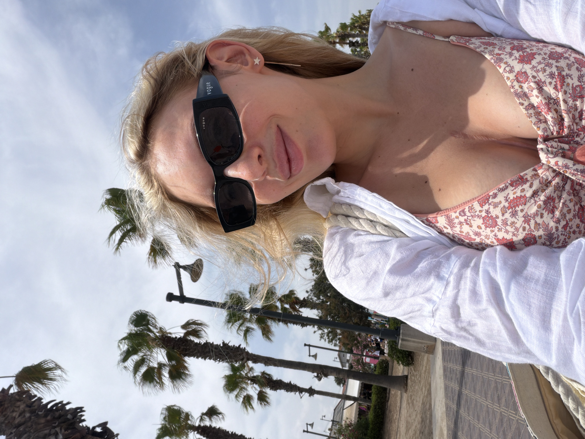 A woman with blonde hair is taking a selfie outdoors. She is wearing sunglasses and a floral bikini top. Her chest is visible, and she has a scar across her sternum. A white shirt is draped over her shoulders. She is holding a bag with a rope strap. In the background, there are palm trees and a cloudy sky. The location appears to be a public walkway or promenade, possibly near a beach or park, with people and buildings in the distance. The lighting suggests it is daytime.