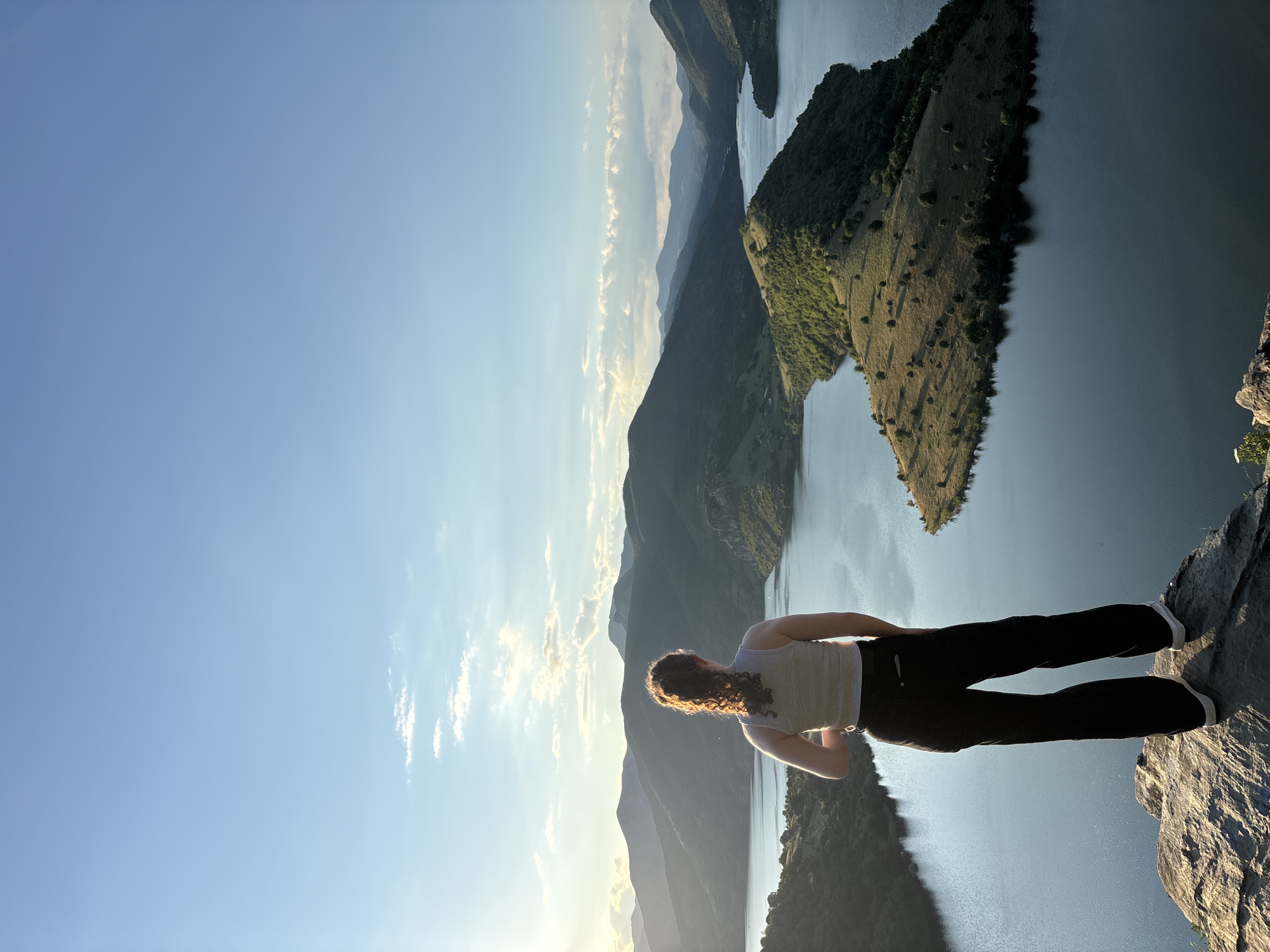 An individual stands on a rocky precipice overlooking a vast, serene lake nestled within a mountainous landscape. The person, viewed from the back, has curly brown hair and wears a light-colored tank top and dark, wide-leg pants. Their posture suggests contemplation as they gaze towards the horizon.
The lake, a deep blue, meanders through a valley dotted with verdant hills and islands. One prominent island, shaped like a spade, is covered in grassy slopes and scattered trees, catching the warm, golden light of what appears to be a sunset or sunrise. Layers of distant, undulating mountains recede into the background, their peaks softened by the atmospheric haze. The sky above is a clear, pale blue, with a few wispy clouds near the horizon. The overall scene evokes a sense of tranquility and natural beauty.
This location is the Studen Kladenets Dam, near the town of Borovitsa, in Bulgaria. The unique land formations and the expansive water body are characteristic of the Eastern Rhodope Mountains region.