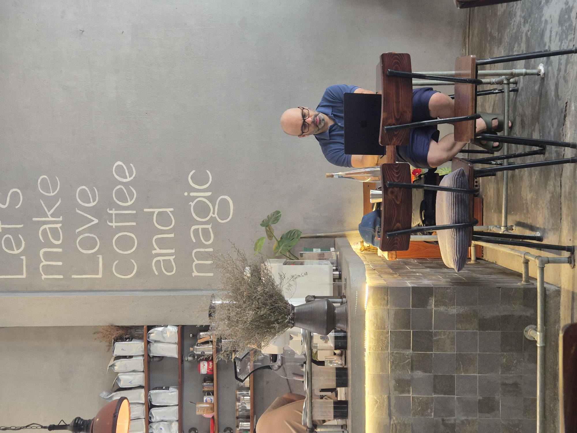 A man with a bald head and glasses sits at a small wooden table, looking at a black laptop. He is wearing a blue polo shirt and blue shorts, and green sandals. The man is seated in a cafe in Da Nang, Vietnam, as indicated by the inscription "Let's make love coffee and magic" on the wall. Behind him, a coffee bar is visible with various coffee-making equipment, bags of coffee, and a vase of dried flowers. The cafe has a modern, industrial aesthetic with exposed pipes and concrete walls. The lighting is warm and inviting, with a string of lights above the counter.