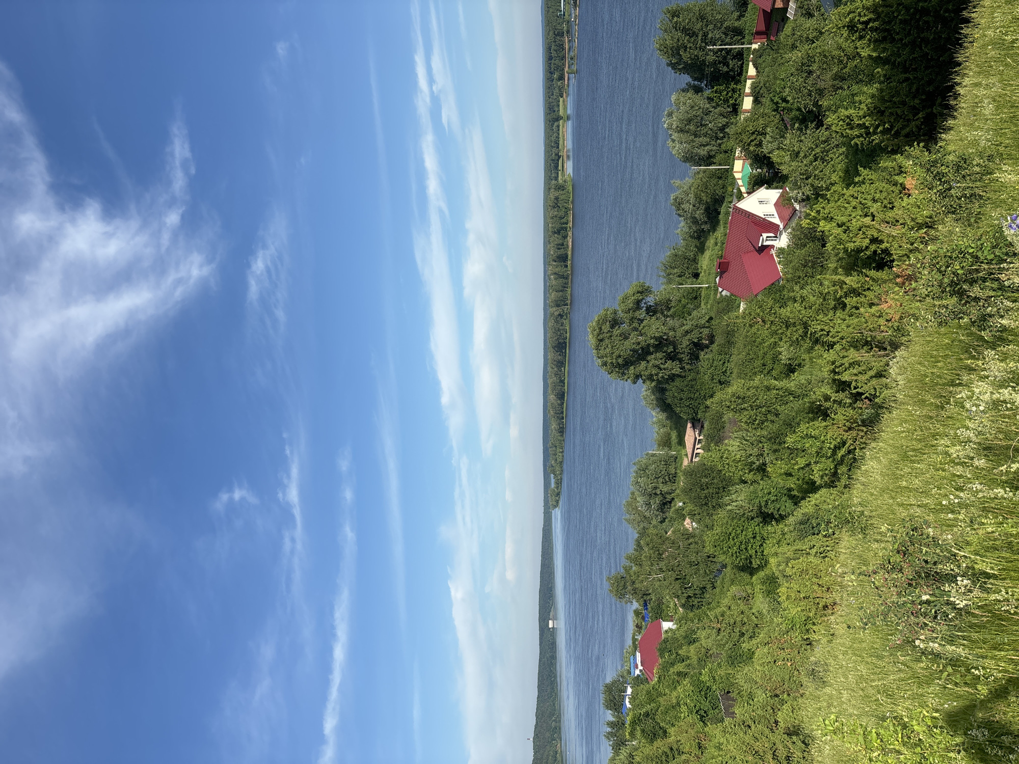 This is an eye-level, wide-angle, outdoor, full shot of a panoramic river landscape. The scene is dominated by a wide, calm river that stretches across the frame, meeting a distant, tree-lined horizon. The sky above is a bright blue with scattered wispy white clouds. In the foreground, a lush green hillside covered in tall grass slopes down towards the river. Dotted amongst the trees on the hillside are several houses with red roofs. The overall impression is one of a peaceful, rural setting. The location is the city of Olkhovka, Russia.