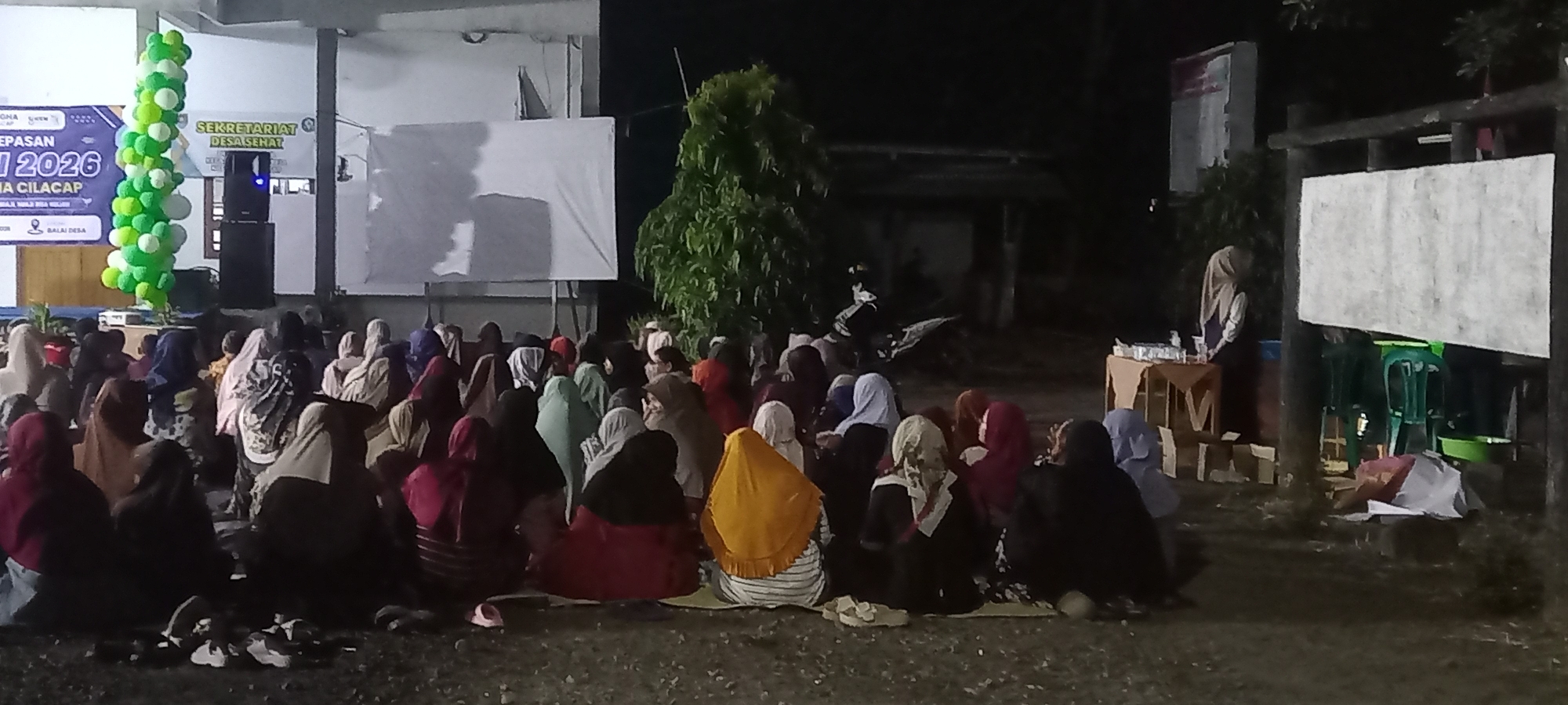 The image depicts an outdoor gathering at night in what appears to be a public space in Sidamulya, Indonesia. A large group of people, predominantly women wearing headscarves, are seated on the ground facing a stage area. The stage features a banner that reads "DIES NATALIS" and "SEKRETARIAT DESA SEHAT". A tall, green and white balloon arch stands to the left of the stage. There is a speaker on a stand and a large blank projection screen. To the right of the stage, there are some tables with items on them and green plastic chairs. The environment is dimly lit, with trees and buildings visible in the background. The ground is covered with gravel. The scene suggests an event or meeting is taking place in a community setting.