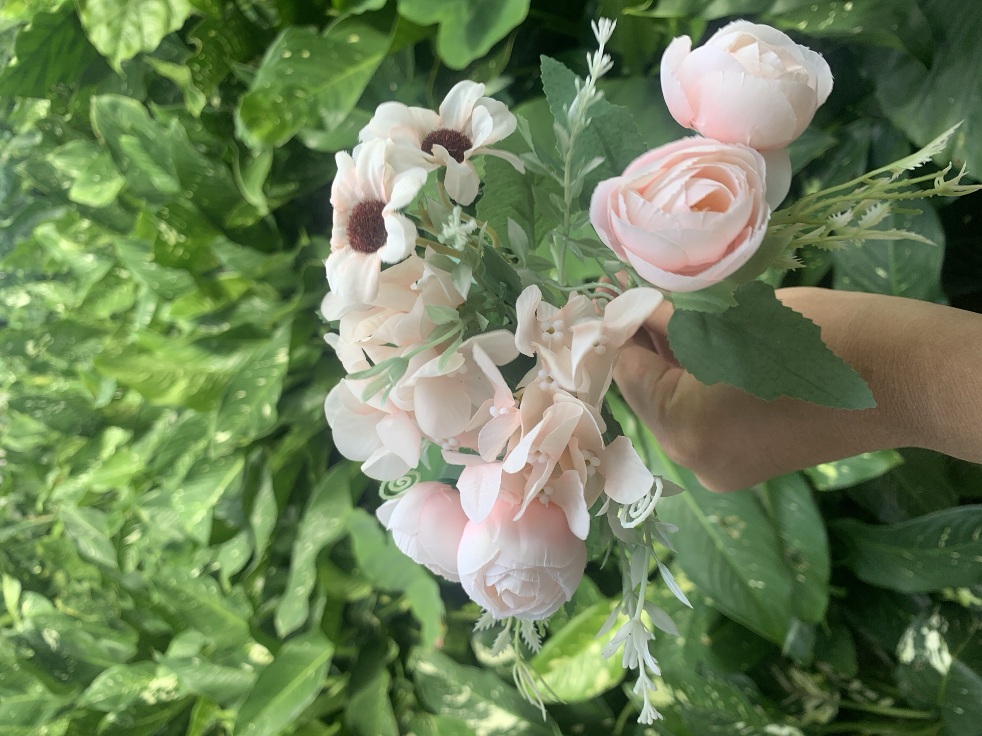 A hand holds a bouquet of light pink artificial flowers in front of a backdrop of lush green foliage. The bouquet features a variety of flower types, including what appear to be roses and daisies. The foliage is dense and appears to be part of an outdoor plant or hedge. The lighting suggests it is daytime. There is no visible indication of the specific location within Kuala Pembuang, Indonesia.