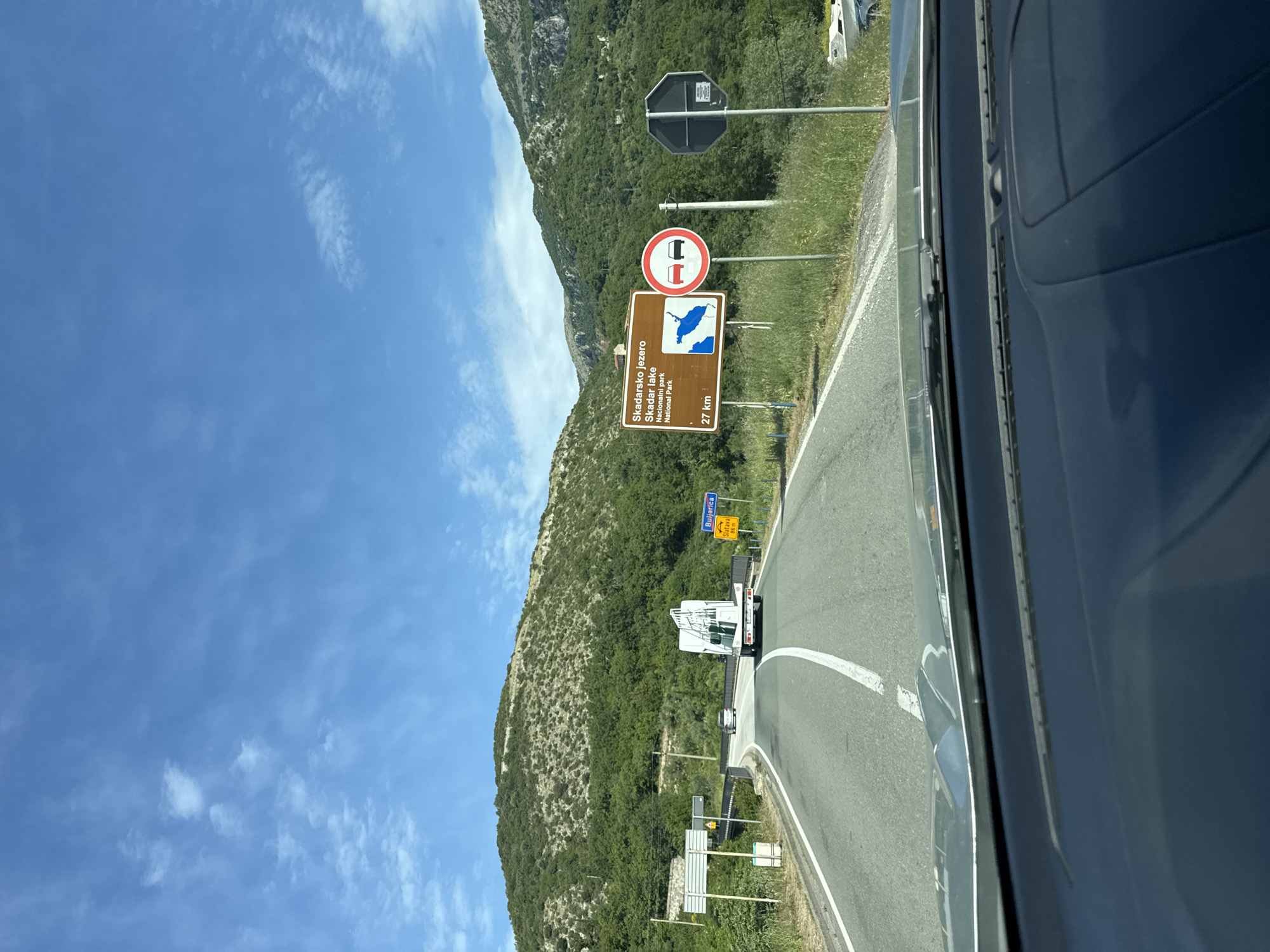The image, taken from inside a vehicle, shows a road winding through a mountainous, green landscape under a blue sky with scattered clouds, which is located on a road in Kaluđerac, Montenegro. Roadside signage indicates that Skadar Lake National Park is 27 kilometers away. The signage also states that Buljarica is 86 meters away, on Slatava Road. Other road signs are visible, including one indicating a restriction for vehicles of a certain size. A white truck is driving on the road ahead. The windshield and side of the vehicle obscure part of the view.