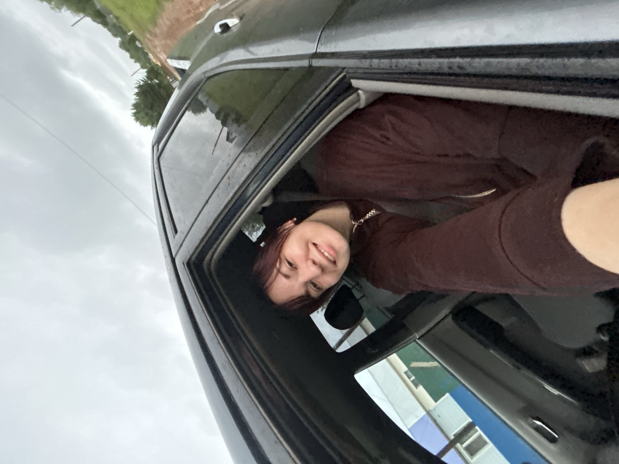 A young woman with dark reddish-brown hair and a light complexion is taking a selfie from inside a dark gray car. She is leaning out of the open passenger window, smiling at the camera. She is wearing a dark reddish-brown hooded sweatshirt with a zipper. The interior of the car is dark.
The scene outside the car appears to be a cloudy day, with a gray sky and some trees visible in the background. The ground appears to be wet, suggesting it may have rained recently. There are some structures visible in the distance, including what looks like buildings with light-colored siding. The image is oriented sideways, with the car door frame running vertically.
The woman's expression is cheerful and engaging. The overall mood of the photo is casual and lighthearted, as if she is enjoying a car ride on a cloudy day. No visible text is present in the image.