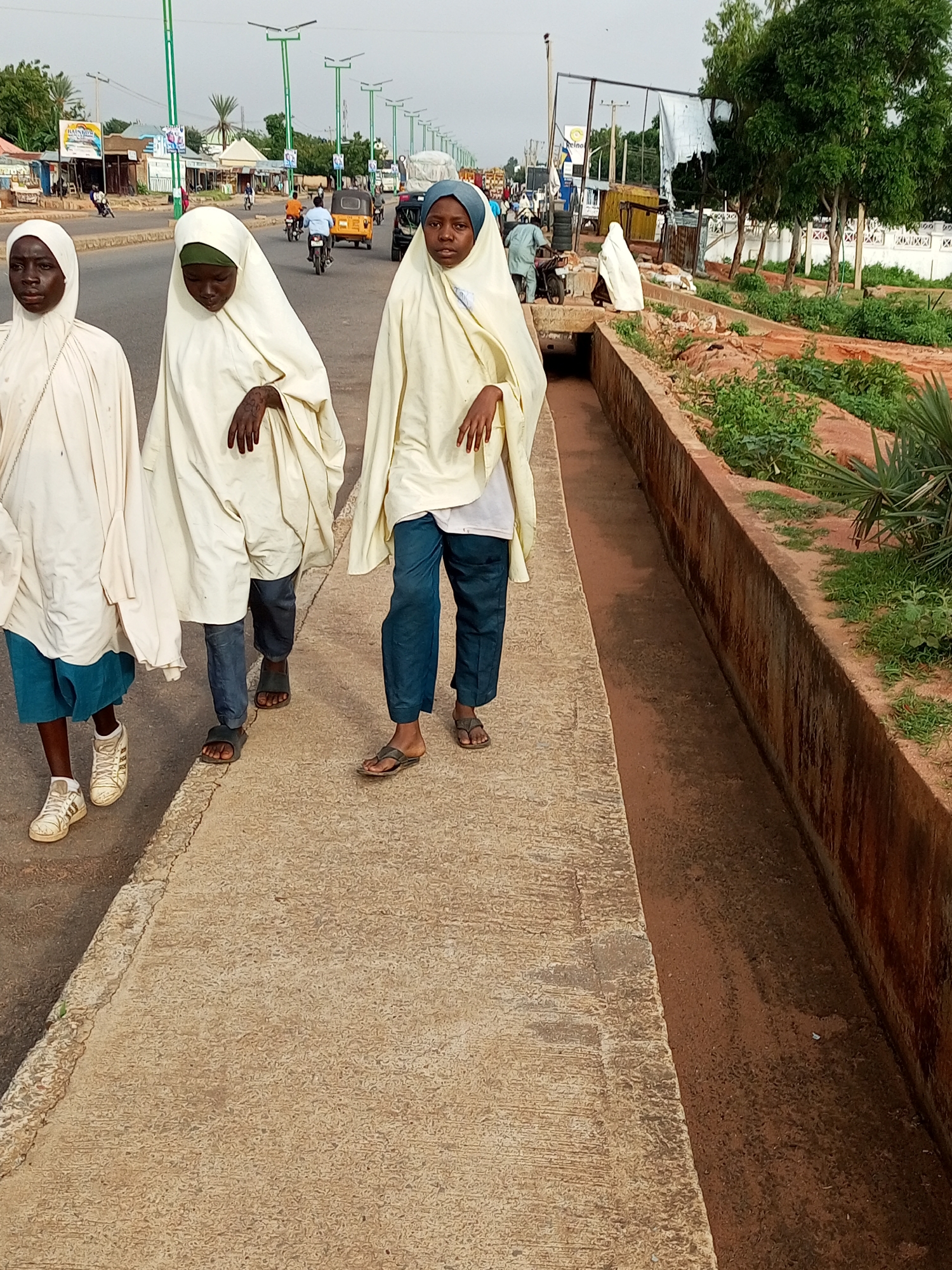 In Garko, Nigeria, three young girls walk along a concrete sidewalk adjacent to a dusty road. The girls are dressed in traditional Islamic attire, with two wearing light-colored hijabs and one in a blue hijab. They are walking in the same direction, with the girl on the left wearing white sneakers and blue shorts visible beneath her robe, and the other two wearing sandals and dark pants. Behind them, a street stretches into the distance, lined with buildings, trees, and a drainage ditch filled with reddish-brown dirt and sparse vegetation. Traffic, including motorcycles and a small yellow vehicle, is visible on the road. The sky is overcast.