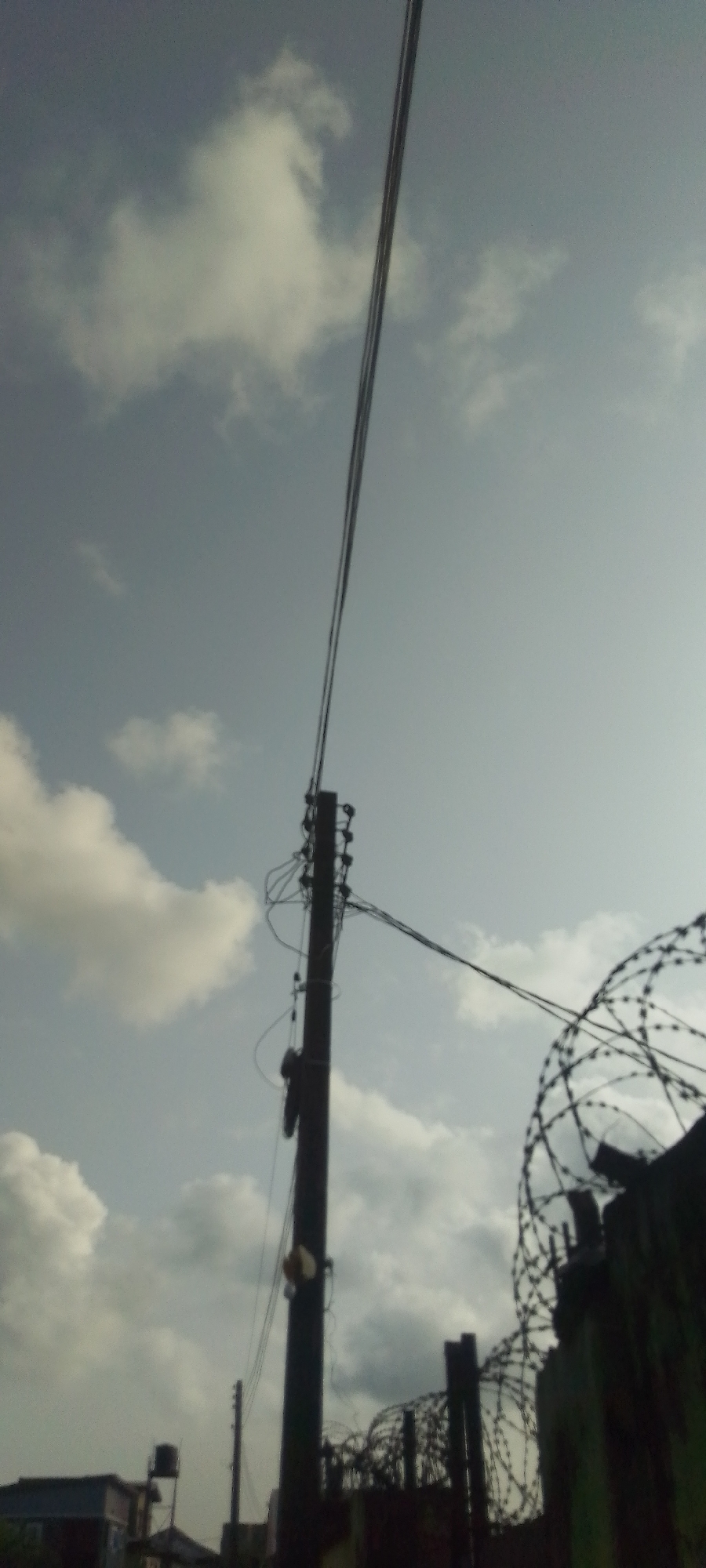 The image shows an urban scene. A utility pole with multiple wires extending from it is centrally positioned. To the right of the pole is a fence topped with barbed wire. The sky is overcast with scattered clouds. Buildings and a water tower are visible in the background. Location: Unknown, Unknown.