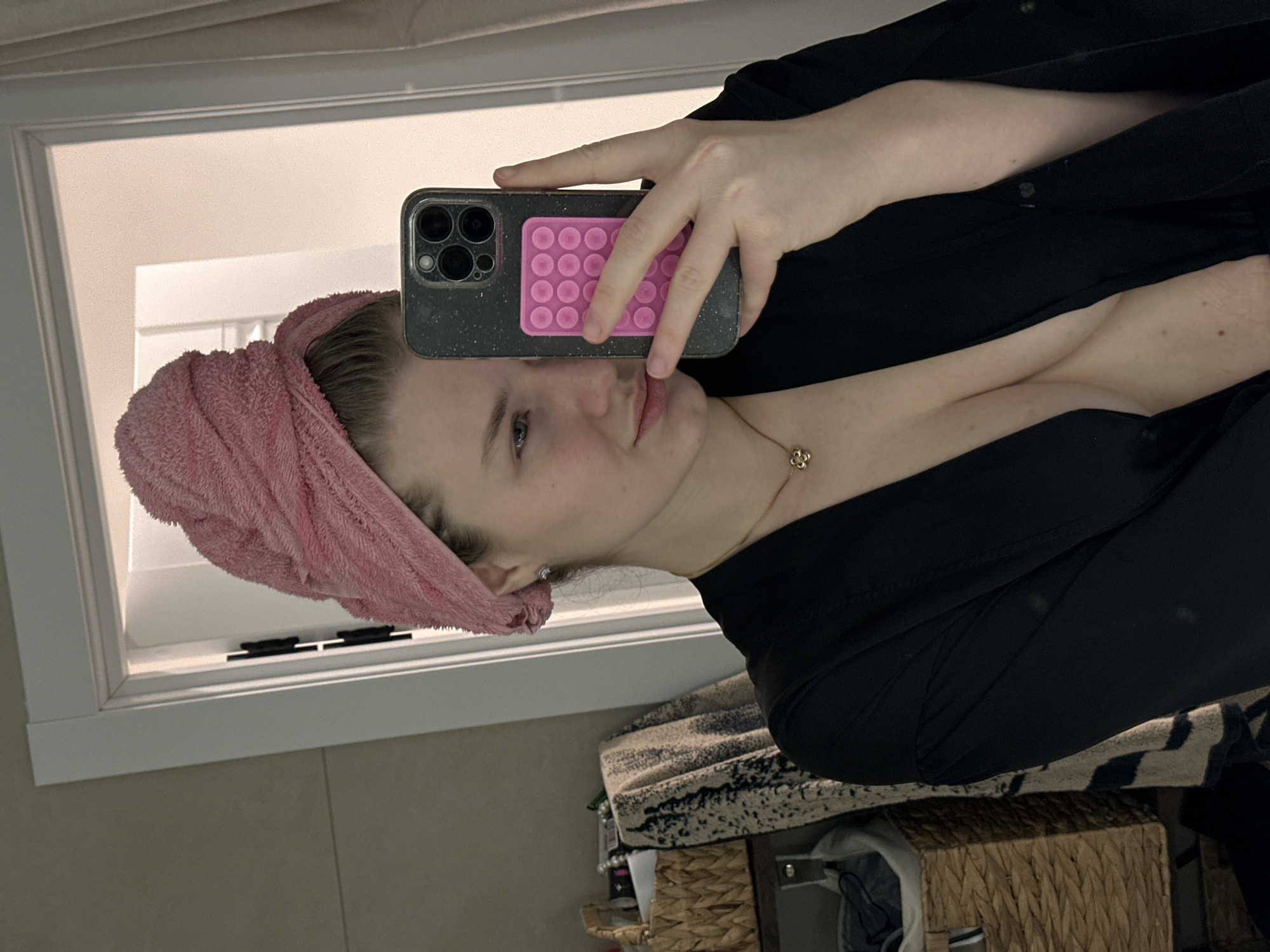 The image shows a woman taking a selfie in a bathroom mirror. She has a pink towel wrapped around her head and is wearing a black robe. She is holding a phone with a pink pop socket. There are bathroom cabinets, towels and other objects around the edges of the shot. The location, Печищи, Russia, cannot be determined from the image content.