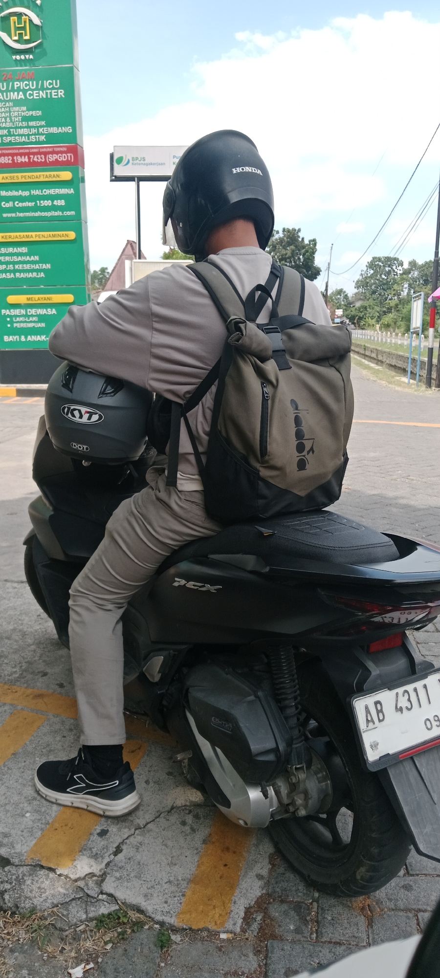 In Maguwoharjo, Indonesia, a person sits on a black Honda PCX scooter, facing away from the camera. They are wearing a grey long-sleeved shirt, beige pants, and black athletic shoes with white accents. A black helmet with "HONDA" written on the back is on their head, and another dark grey helmet with "KYT" and "DOT" logos is held in their lap. A khaki green backpack with black straps and black trim is worn on their back, featuring a "diadora" logo on the front. The scooter's license plate reads "AB 4311 Y." The scene takes place on a paved road with yellow markings. In the background, a tall green sign with white text indicates services like "24 JAM U / PICU / ICU TRAUMA CENTER" and "AKSES PENDAFTARAN" with a contact number. A smaller sign with a "BPJS Ketenagakerjaan" logo is also visible. Trees and buildings are present in the distance, under a bright sky with scattered clouds.