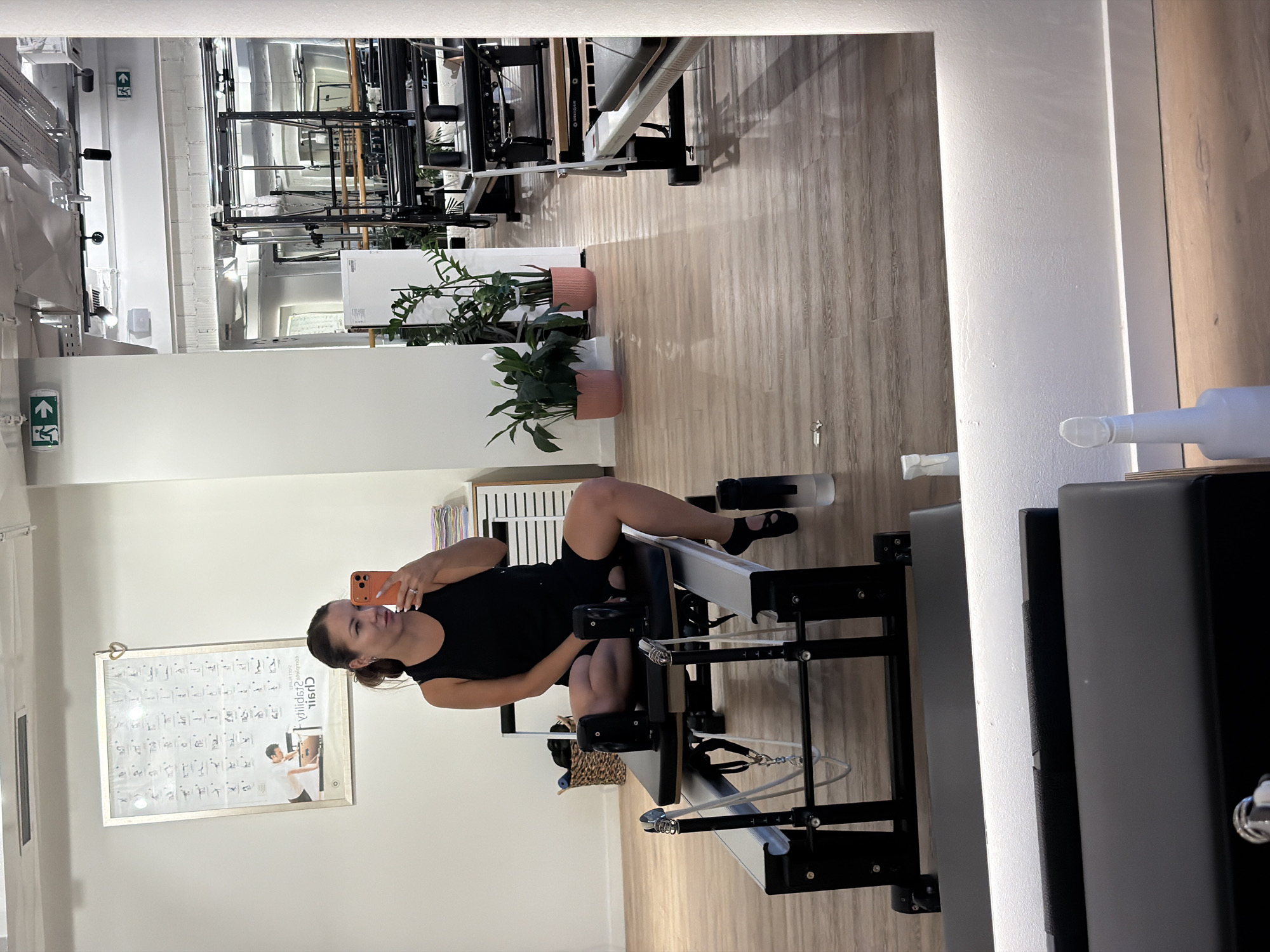 A woman, seen in a reflection in a mirror, sits on a black Pilates reformer machine in a studio. She is wearing a black tank top and black shorts, with black open-toed shoes. She holds an orange smartphone in her right hand, seemingly taking the photo. The studio has light wooden floors and white walls. A poster displaying various Pilates exercises is on the wall behind her, to her left. In the background, other Pilates reformers and equipment are visible. Plants are placed in pink pots on the floor. The scene is brightly lit, with overhead lights and a strip of light below the mirror. The reflection shows a spacious room with large windows or glass doors leading to other areas of the studio, some with more gym equipment. There is a water bottle on the floor next to the woman. The location is a Pilates studio in Boreti, Montenegro.