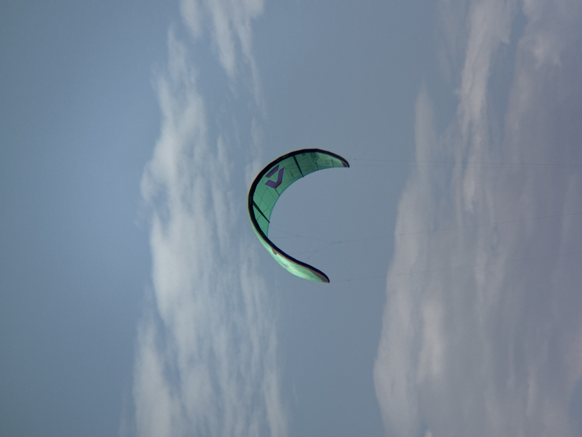A teal kite with a purple logo is suspended in a pale blue sky filled with scattered white clouds. The kite is curved, with its leading edge facing forward and upward, and its trailing edge curving downwards. The kite is a single object in the frame, and there are no people visible. The background consists of the sky and clouds, with no discernible city or geographical features. The image appears to be a close-up shot of the kite against the sky.
